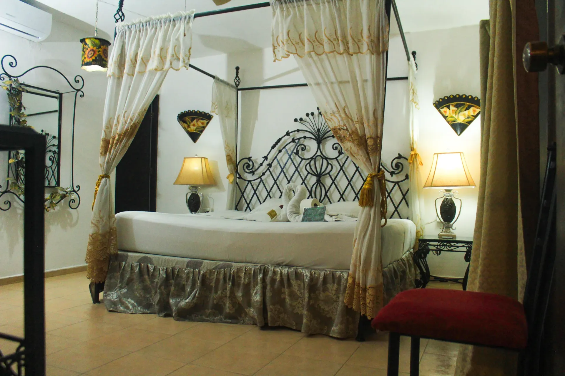 Dormitorio con cama con dosel, cabecera de metal ornamentado, detalles dorados, lámparas y un taburete rojo.