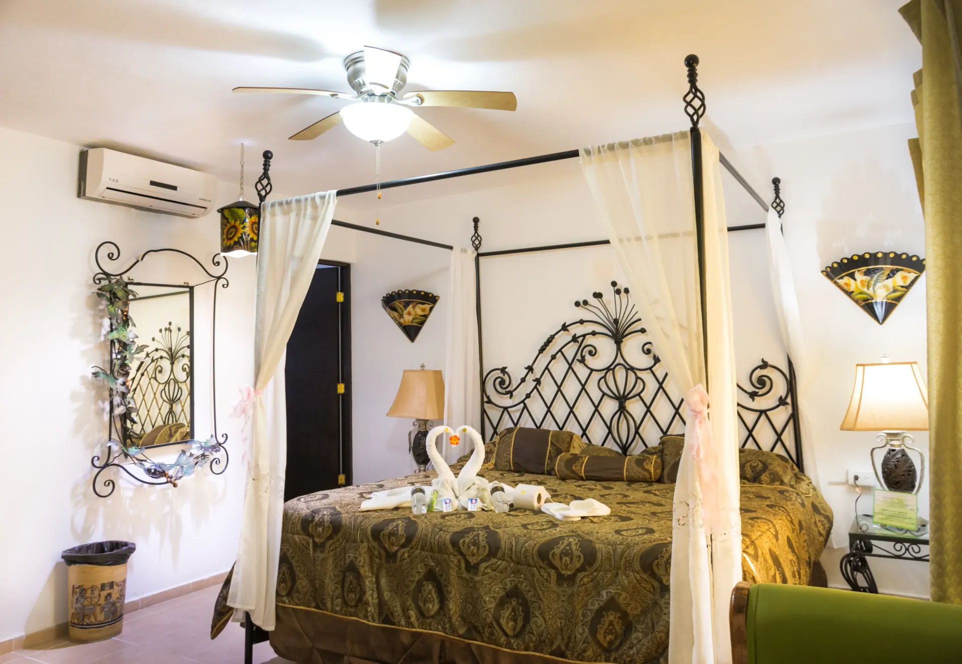 Dormitorio con cama con dosel, cabecera ornamentada, cortinas transparentes, lámparas y espejo decorativo.