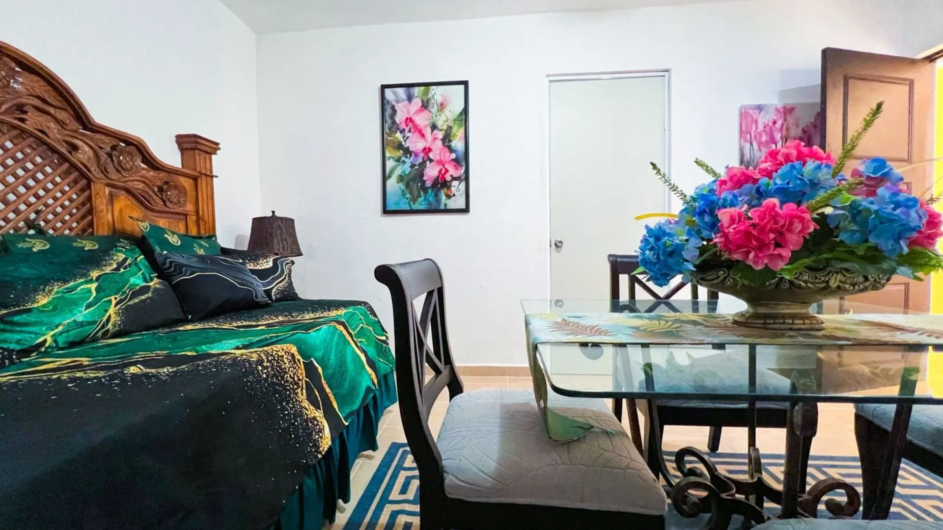 Dormitorio con cama, mesa de comedor y arreglo floral. Colores verde, azul y rosa.