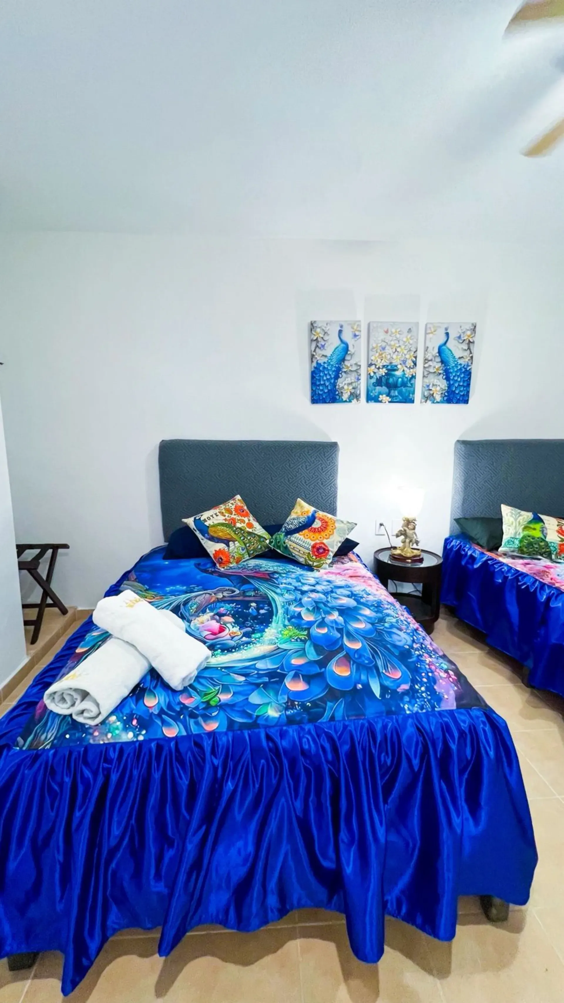 Dos camas individuales con ropa de cama de pavo real azul en un dormitorio con ventilador y arte mural.