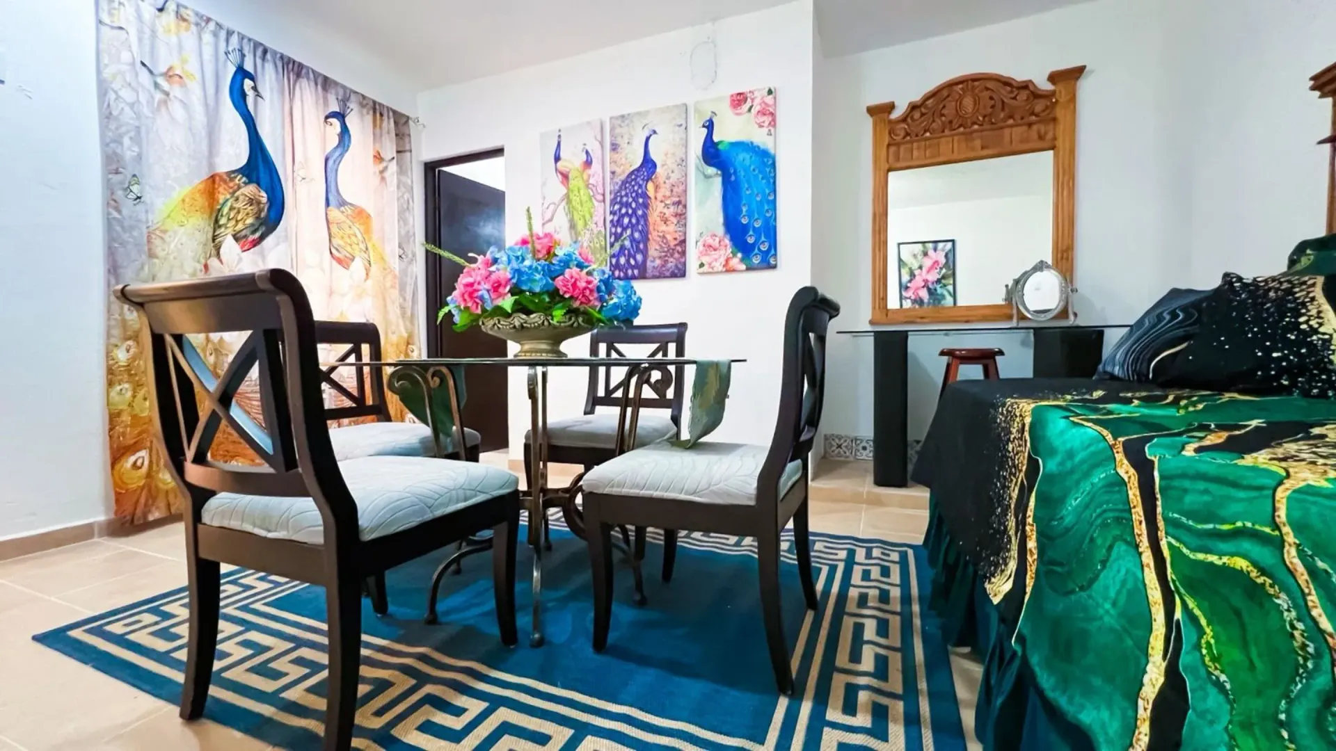Comedor con arte de pavo real, una mesa y sillas sobre una alfombra azul, junto a una cama con ropa de cama verde.