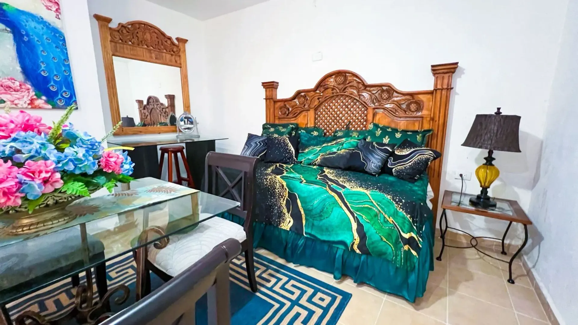 Dormitorio con cabecero de madera tallada, ropa de cama en tonos turquesa y dorado, mesa, espejo, flores y lámpara.