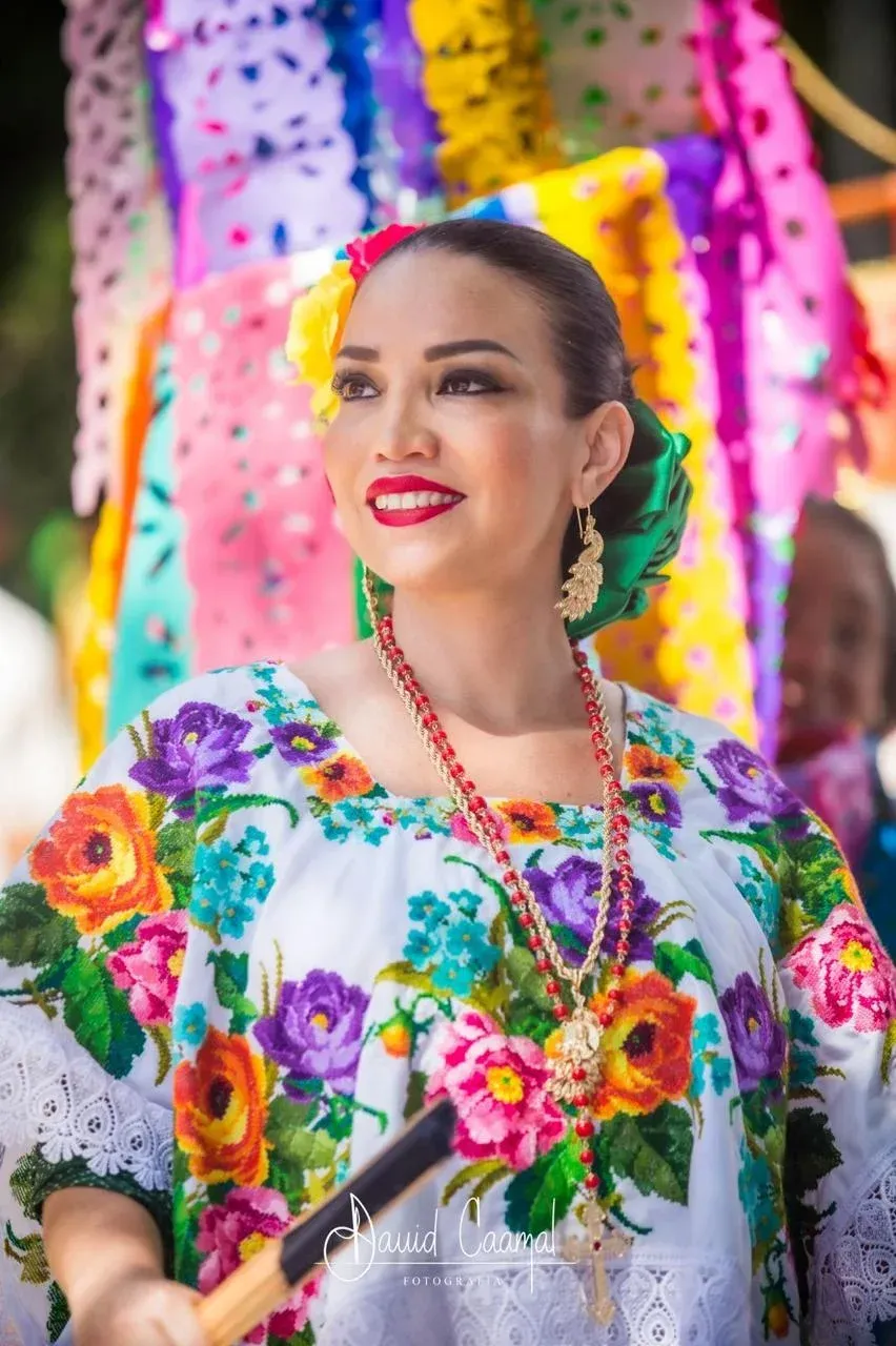 Woman in colorful embroidered dress, smiling, possibly at a festival, with floral decorations.