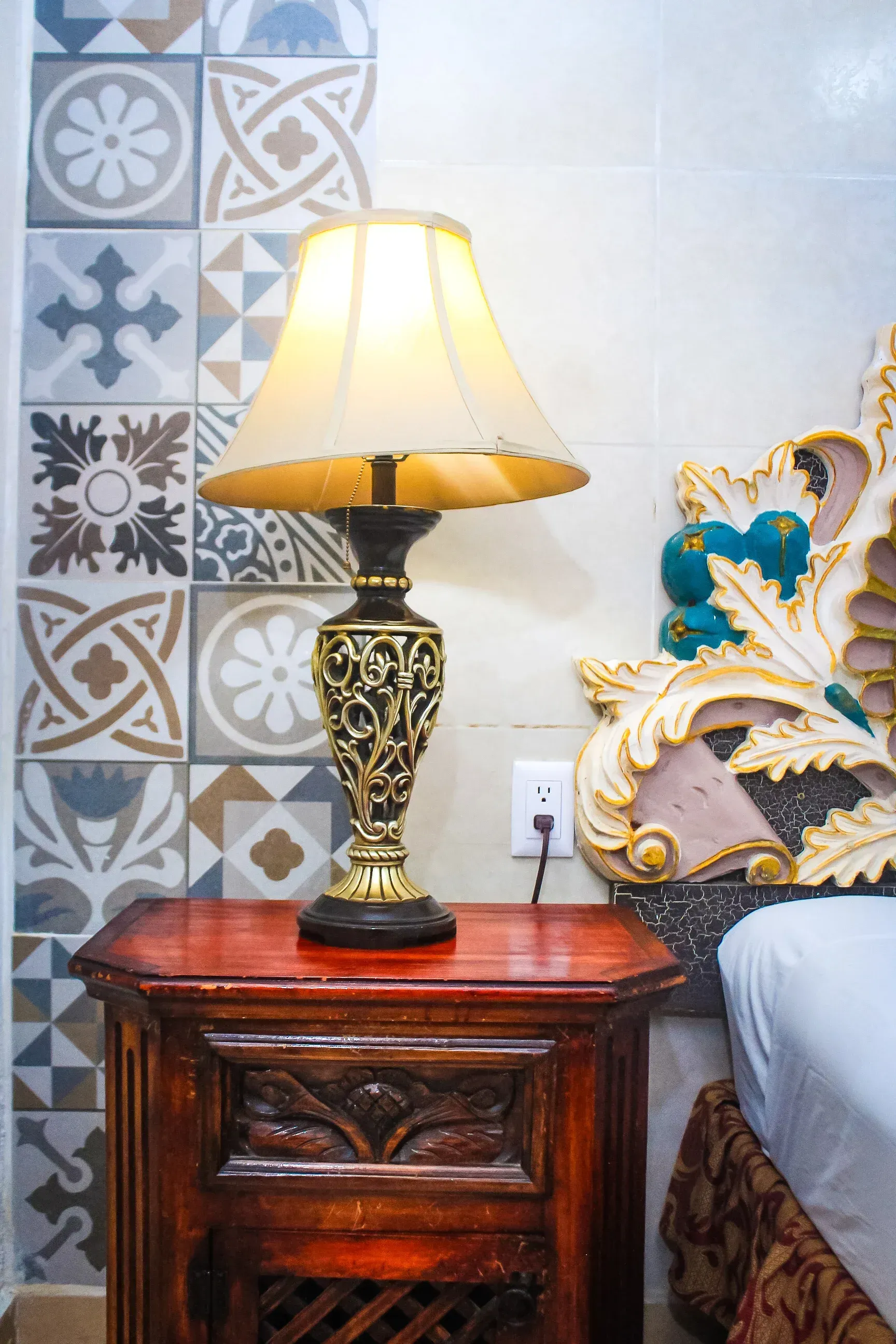 Una mesita de noche con una lámpara, contra una pared estampada, al lado de una cama con un cabecero ornamentado.