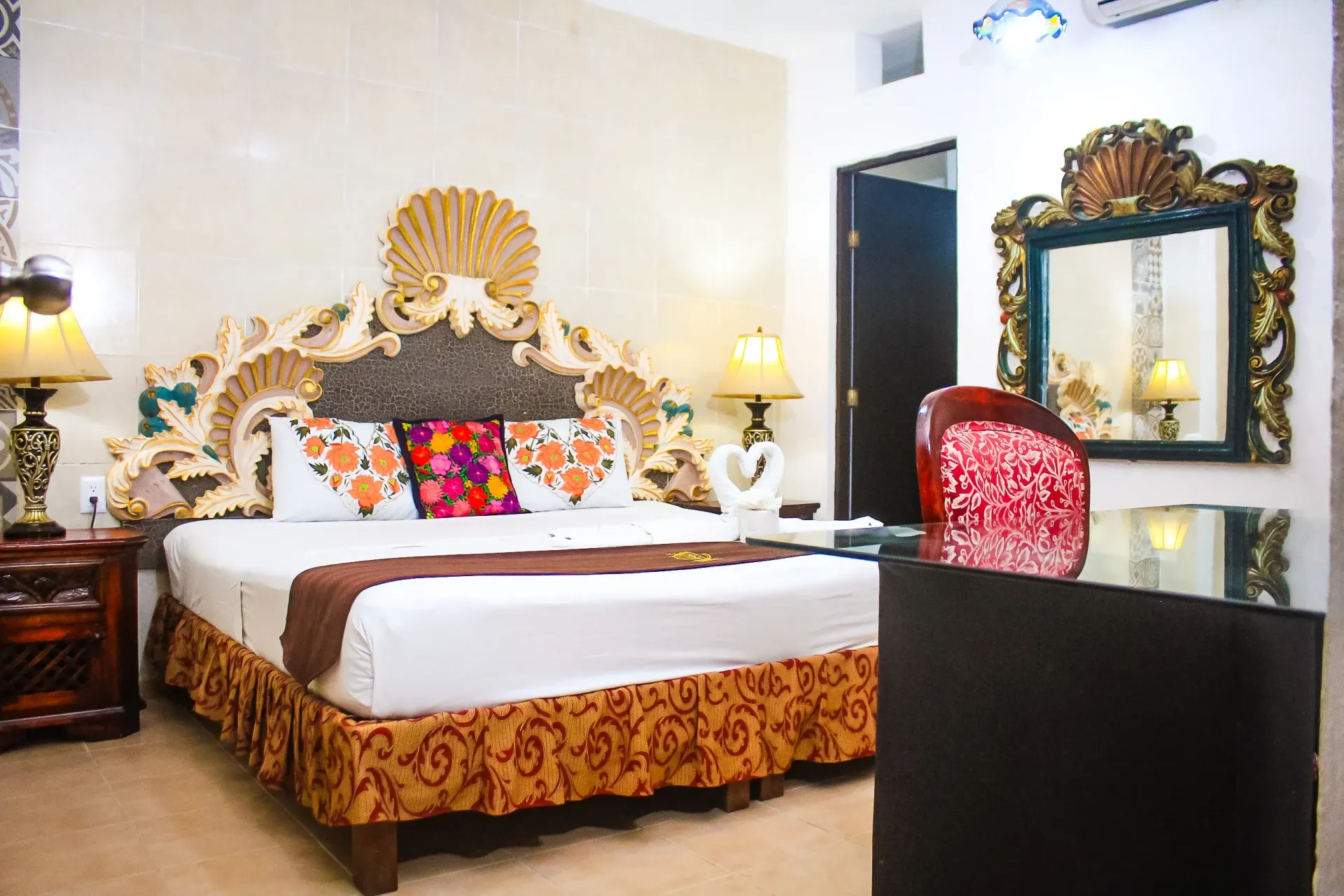 Dormitorio con cabecero ornamentado, colcha, lámparas y espejo decorativo.