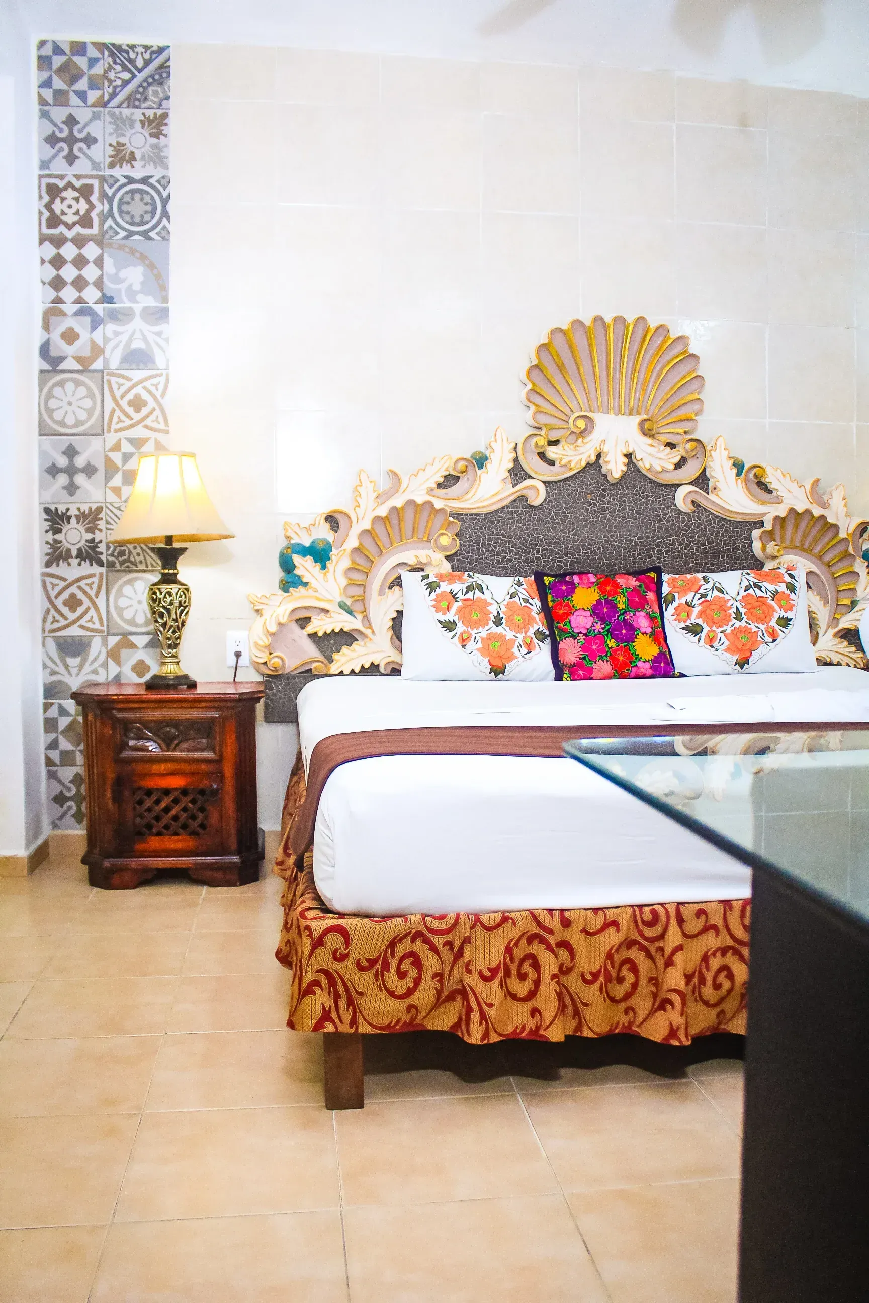 Dormitorio con cabecero ornamentado, mesita de noche y cojines coloridos. Detalles en marrón, beige y estampados.