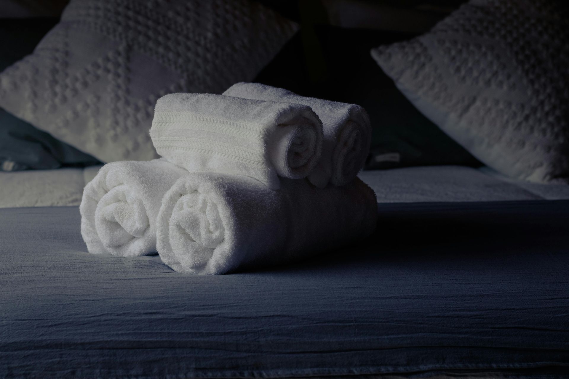 Toallas blancas enrolladas sobre una cama con almohadas decorativas en el fondo.