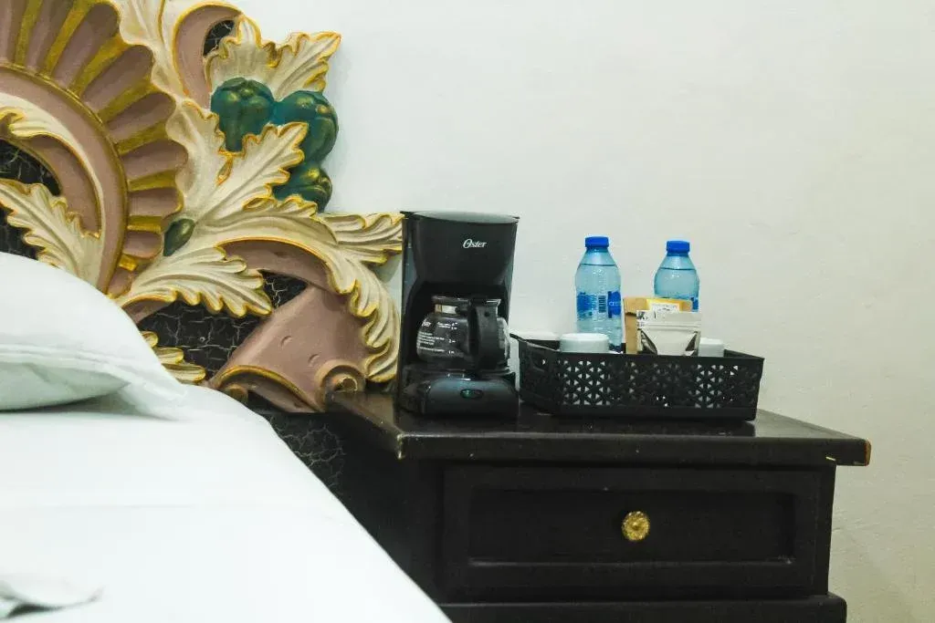 Mesita de noche con cafetera, agua embotellada y tazas junto a una cama con cabecera ornamentada.