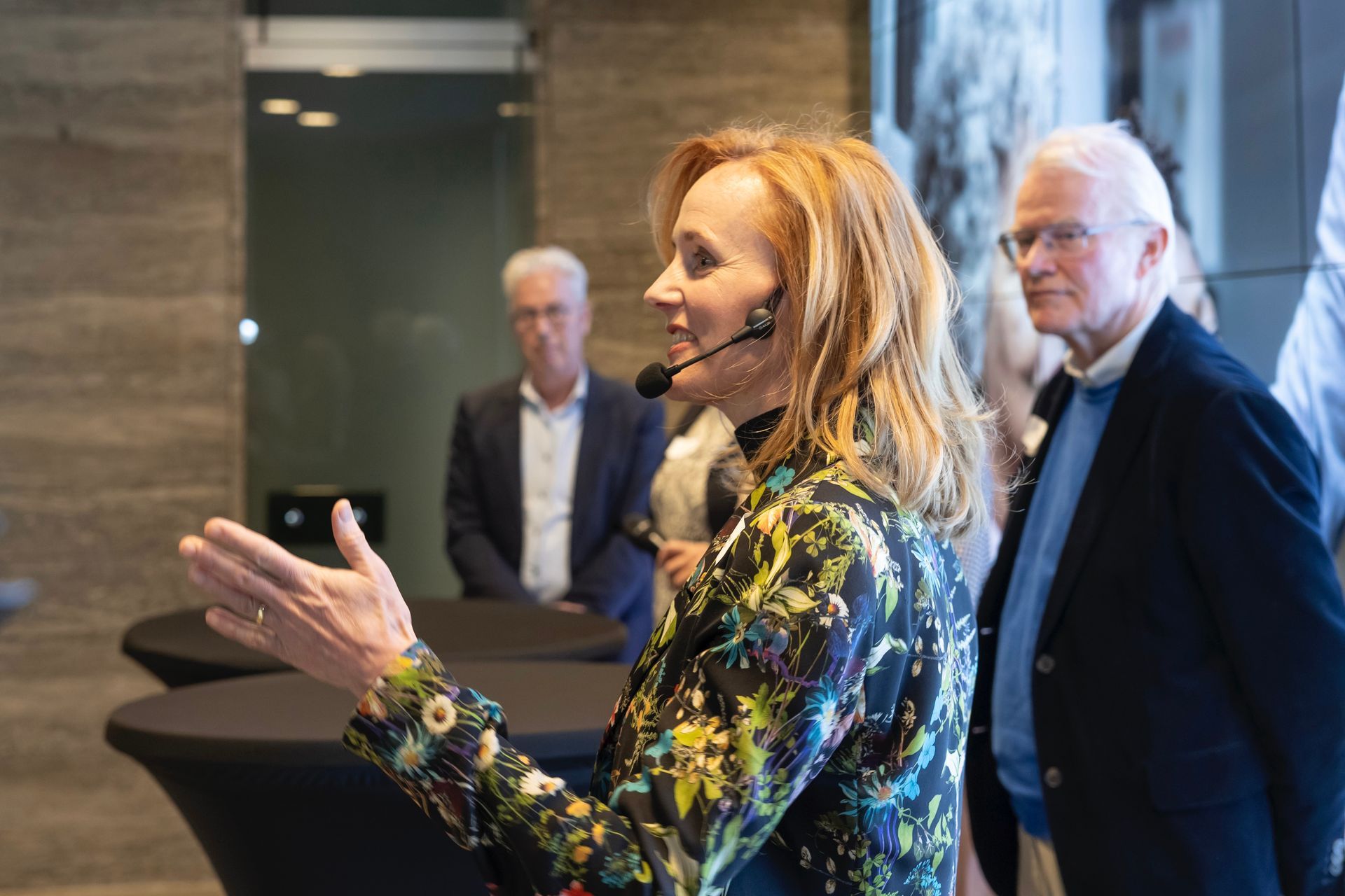 Een vrouw met een microfoon spreekt en gebaart; twee mannen staan ​​achter haar in een modern interieur.