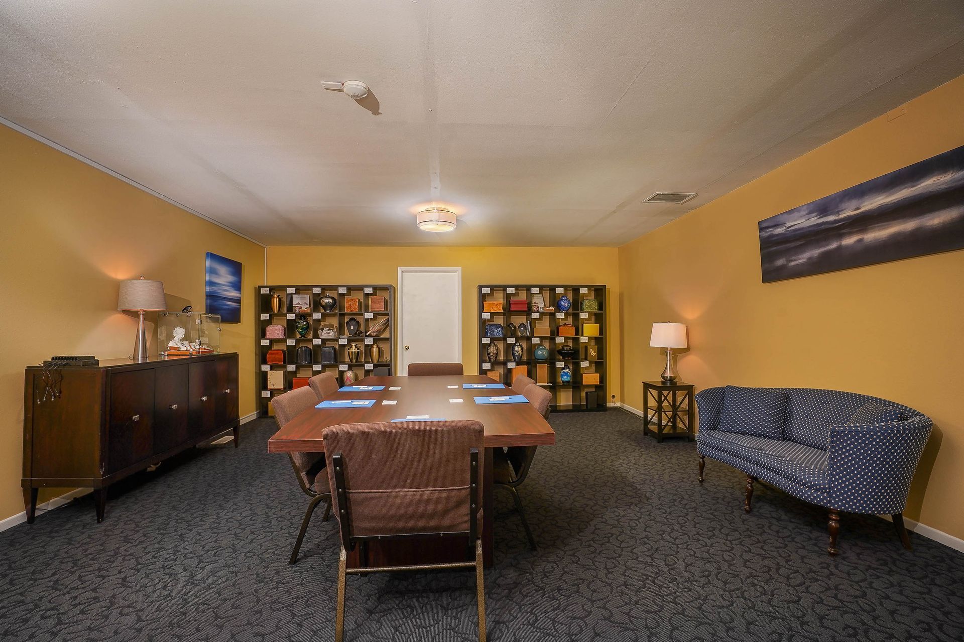 Conference room with a long table, brown chairs, bookshelves, lamps, and a blue sofa on carpet.