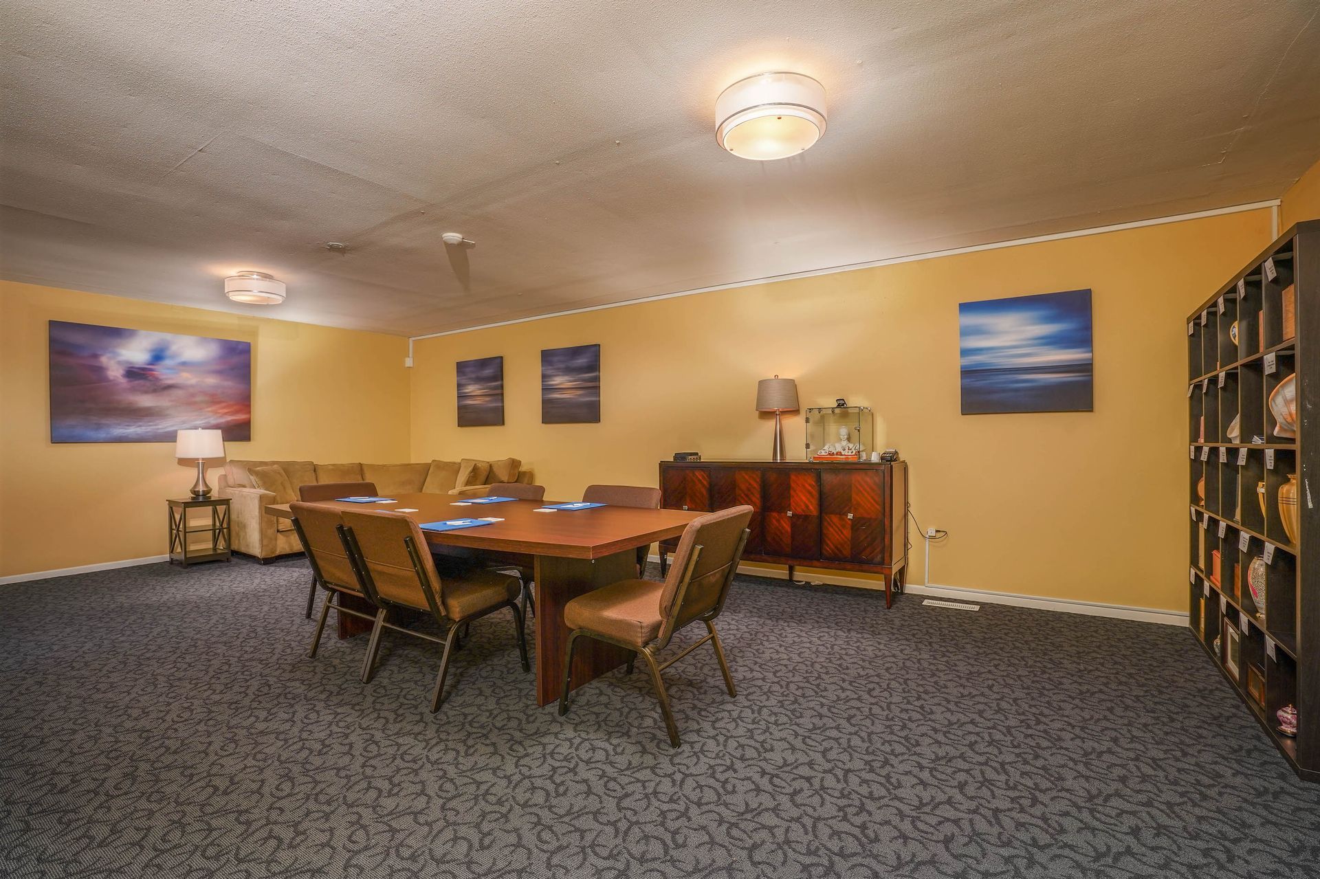 Conference room with wooden table, chairs, sideboard, wall art, and shelving on beige walls and carpet.