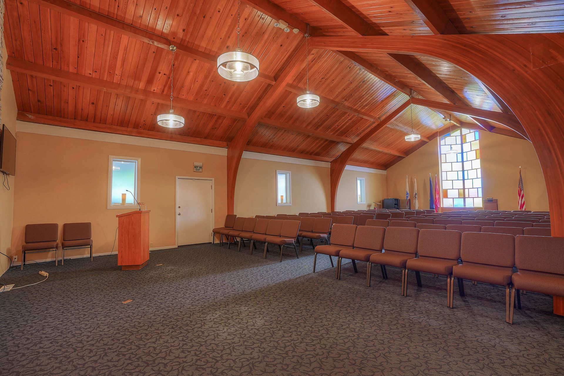 Empty chapel interior with curved wood ceiling, rows of chairs, podium, and stained-glass windows