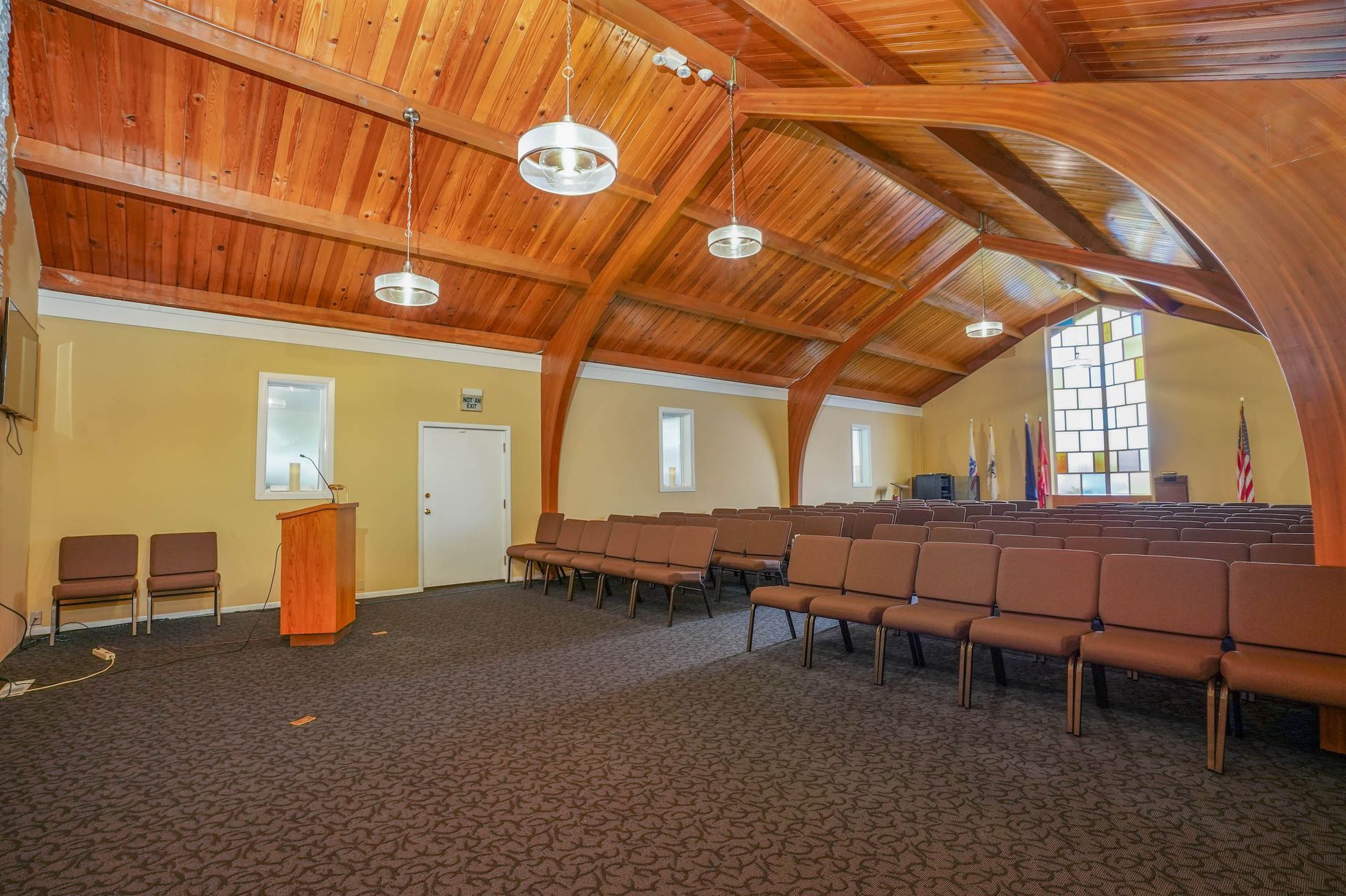 Empty chapel with wooden arched ceiling, rows of brown chairs, a lectern, and stained-glass windows.
