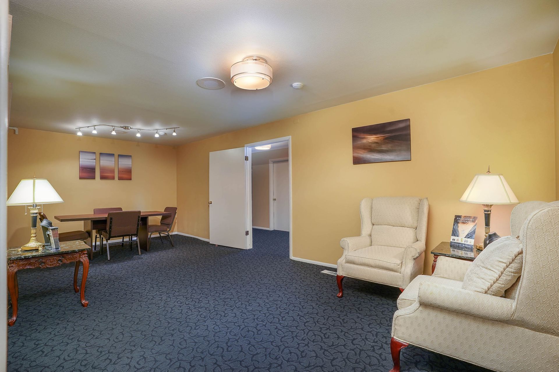 Empty beige lounge with blue carpet, chairs, side tables, lamps, and wall art.