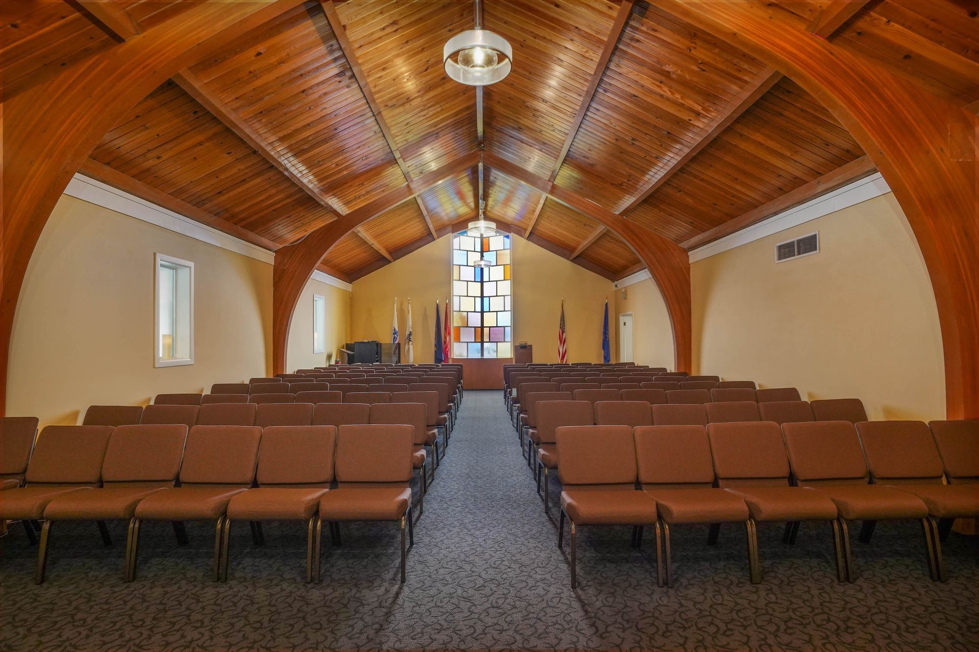 Empty chapel interior with rows of brown chairs, wooden vaulted ceiling, and stained-glass window at the front