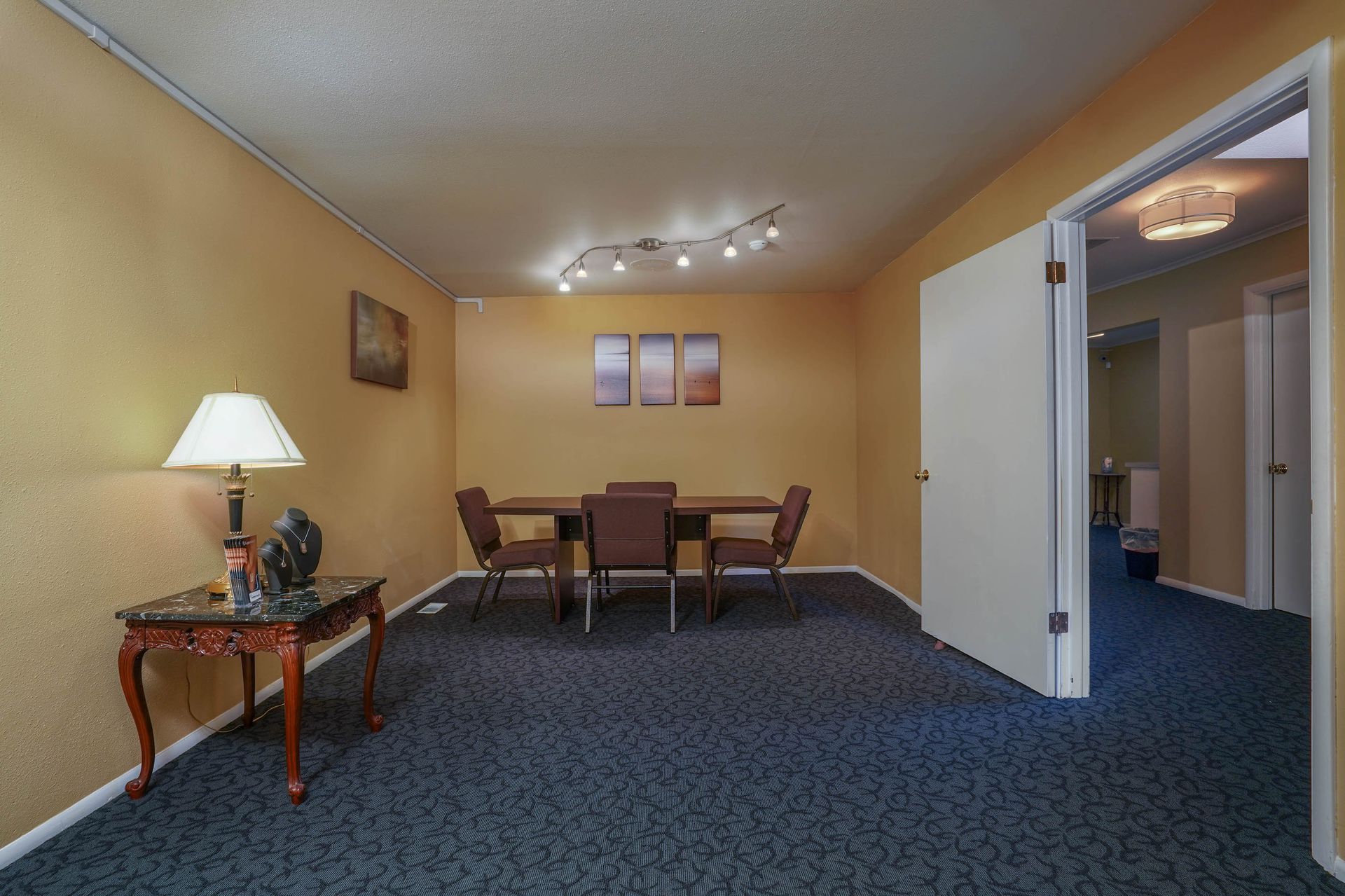 Empty living room with beige walls, blue carpet, dining table, and lamp beside a doorway to the hall.