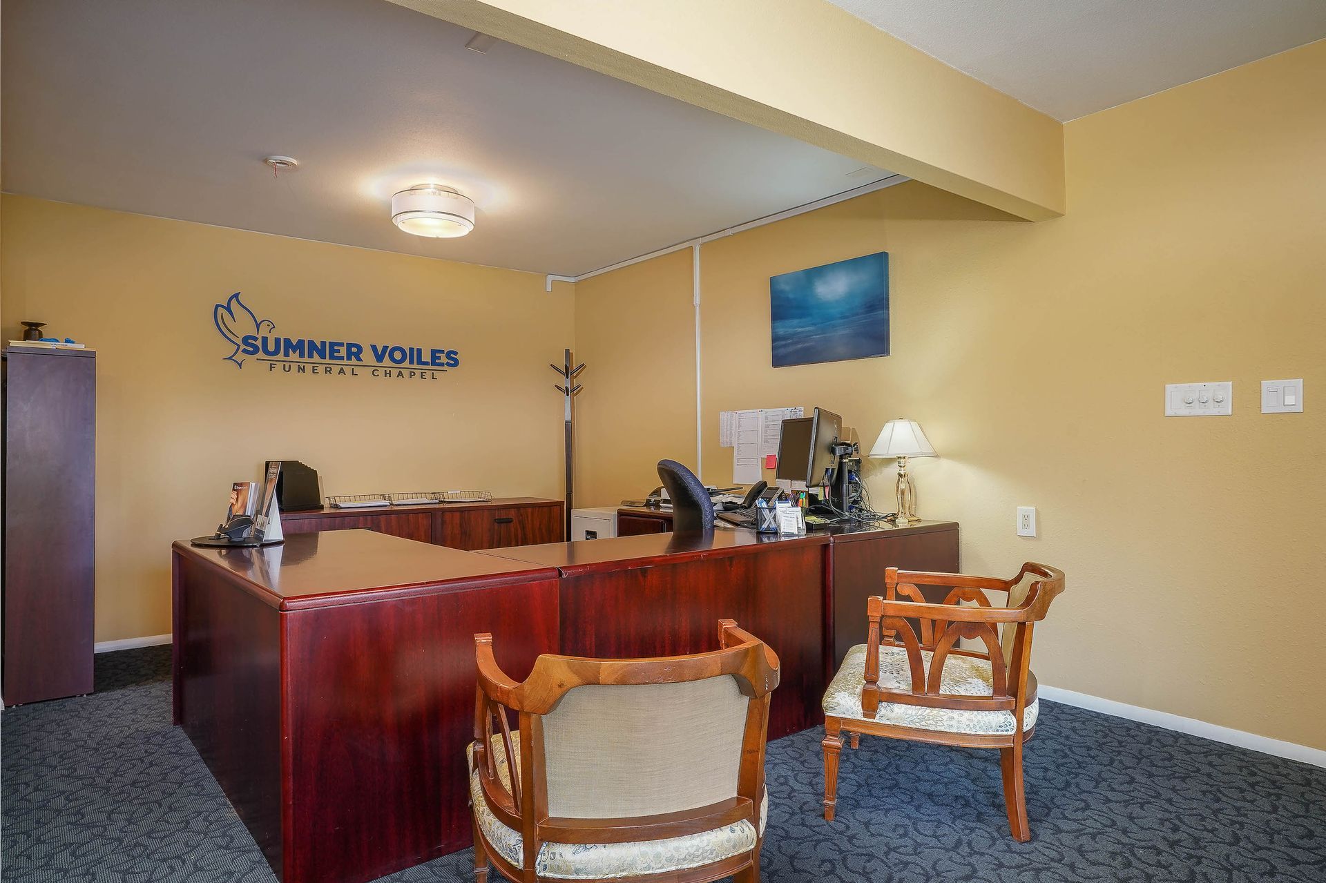 Hotel reception area with dark wood desk, tan walls, and seating chairs, no people visible