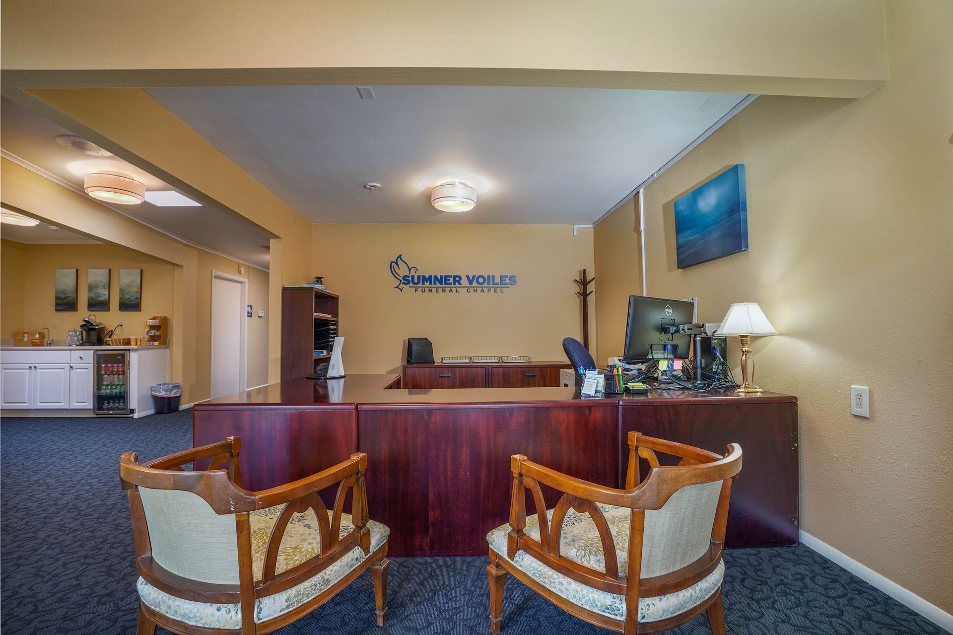 Reception area with wooden desk, two chairs, computer monitor, and wall art in a carpeted lobby