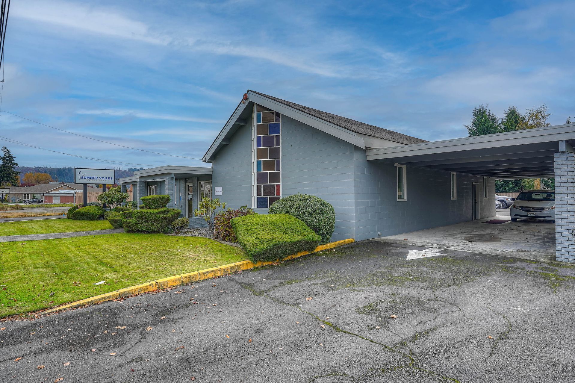 Single-story gray building with carport, wet pavement, and green lawn under a cloudy sky