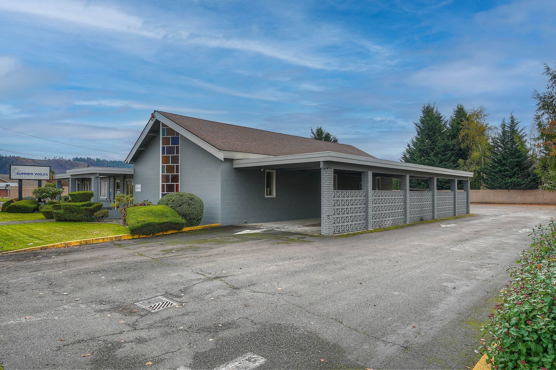 Single-story gray house with a tiled roof, gravel driveway, and covered carport under a cloudy sky