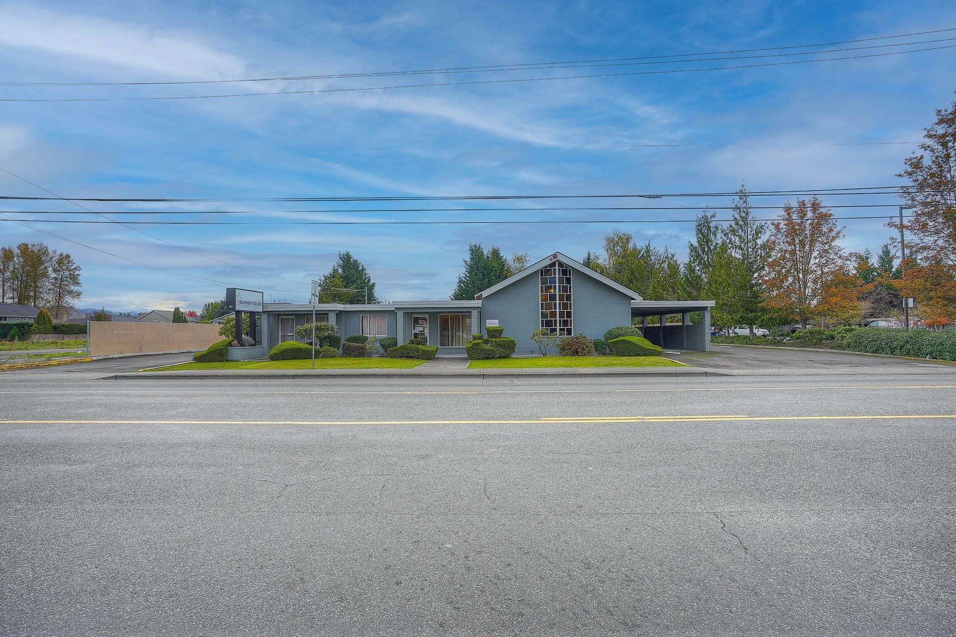 Single-story house with gray roof and blue siding across a wide gravel lot under a cloudy blue sky