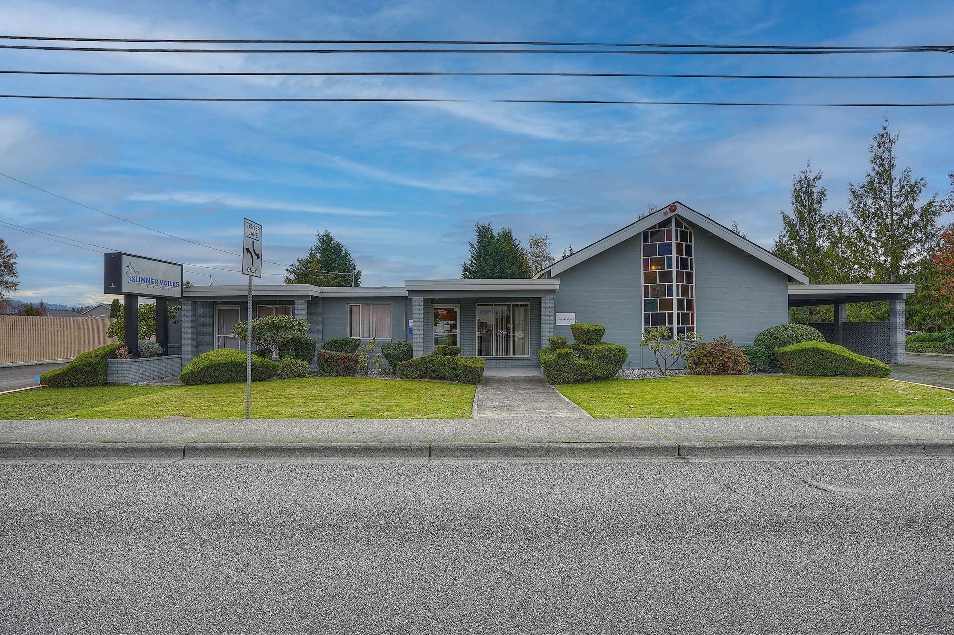 Single-story suburban house with gray roof, green shrubs, and a front sign by the street
