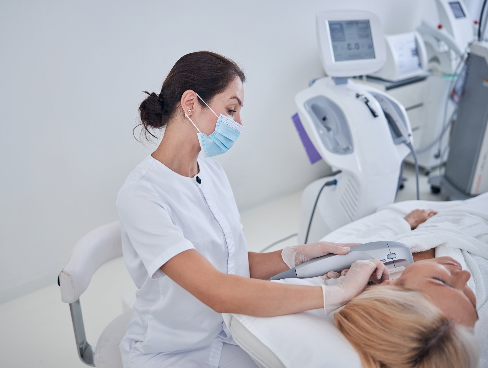 Clinician in white scrubs using an ultrasound probe on a patient in a bright medical exam room