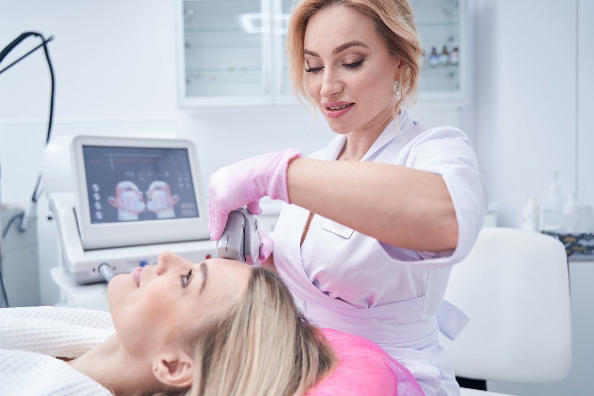 Aesthetic treatment session with a client lying down while a practitioner adjusts a handheld device.