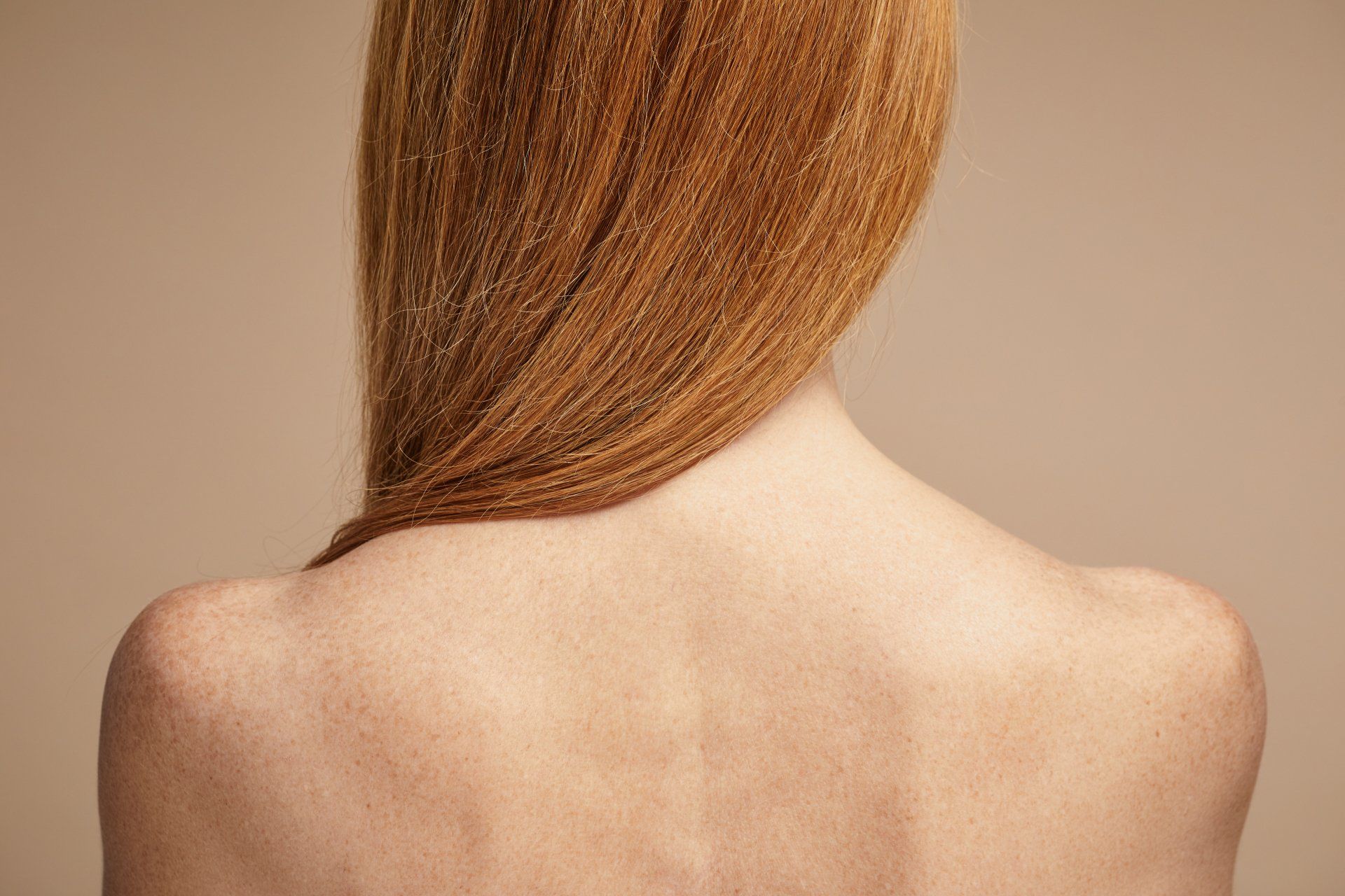 Back view of a person with long red hair draped over bare shoulders against a beige background