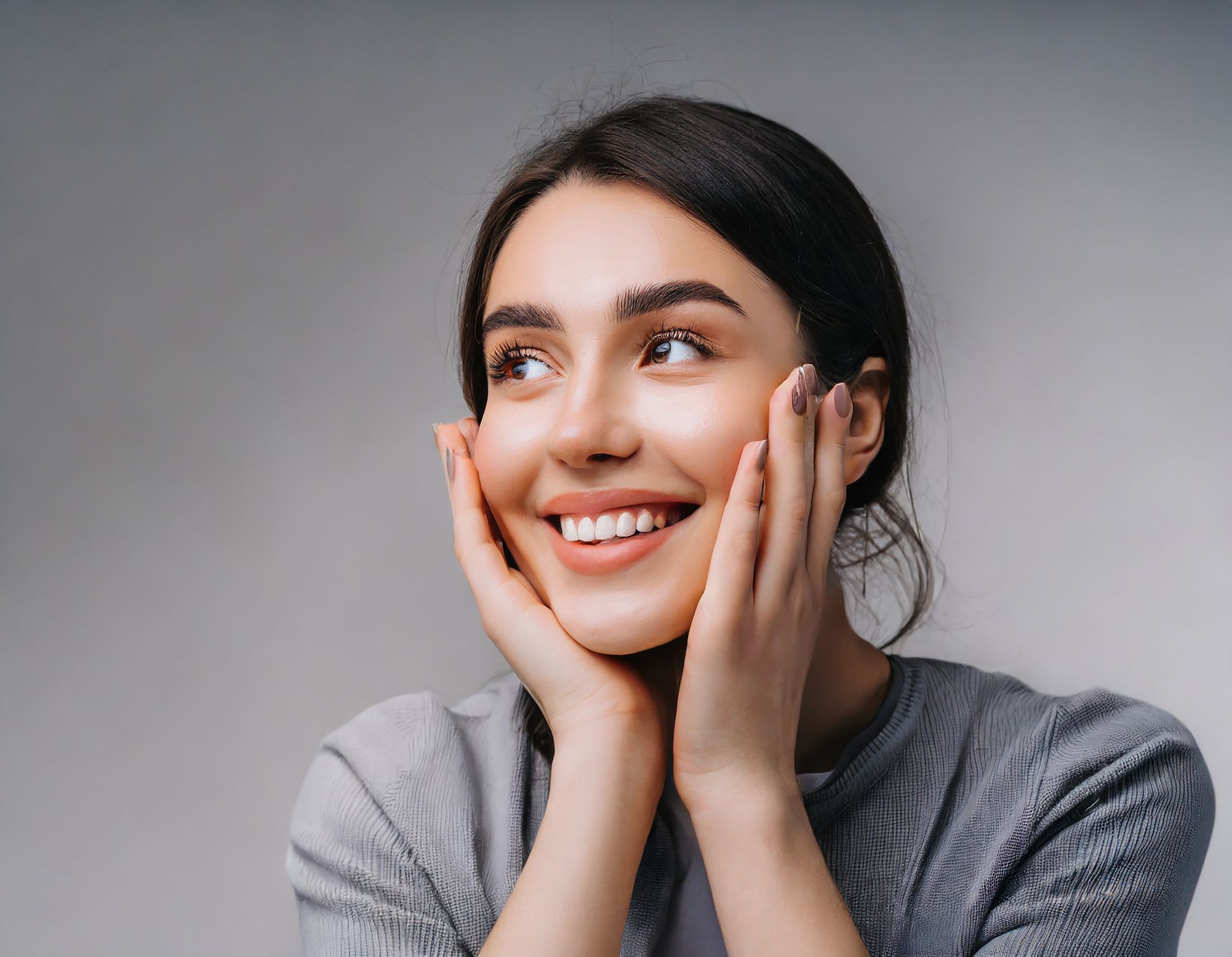 Woman smiling, hands on cheeks, looking up and to the side, against a gray background.