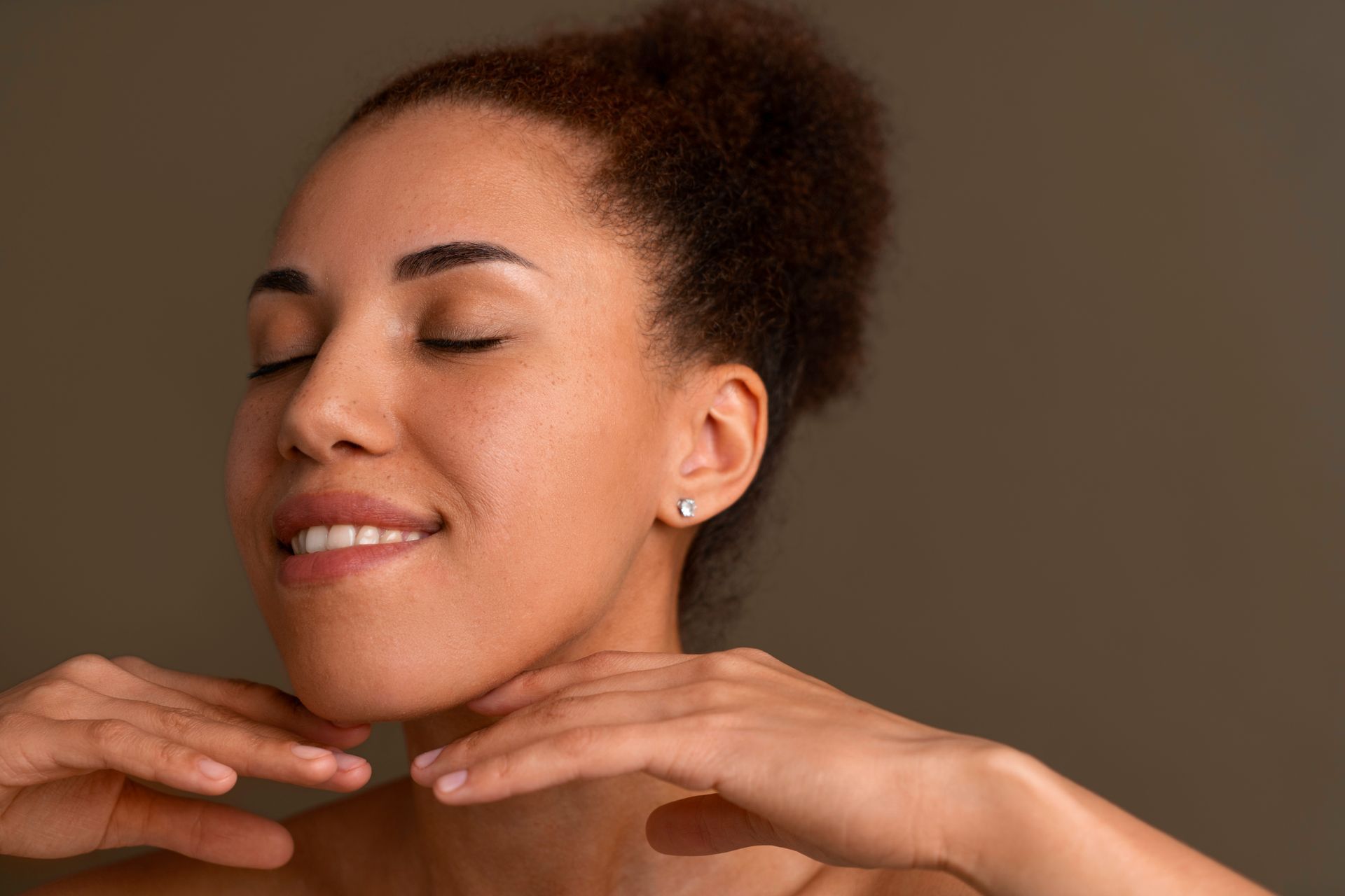 Smiling person with eyes closed, resting chin on hands against a brown background