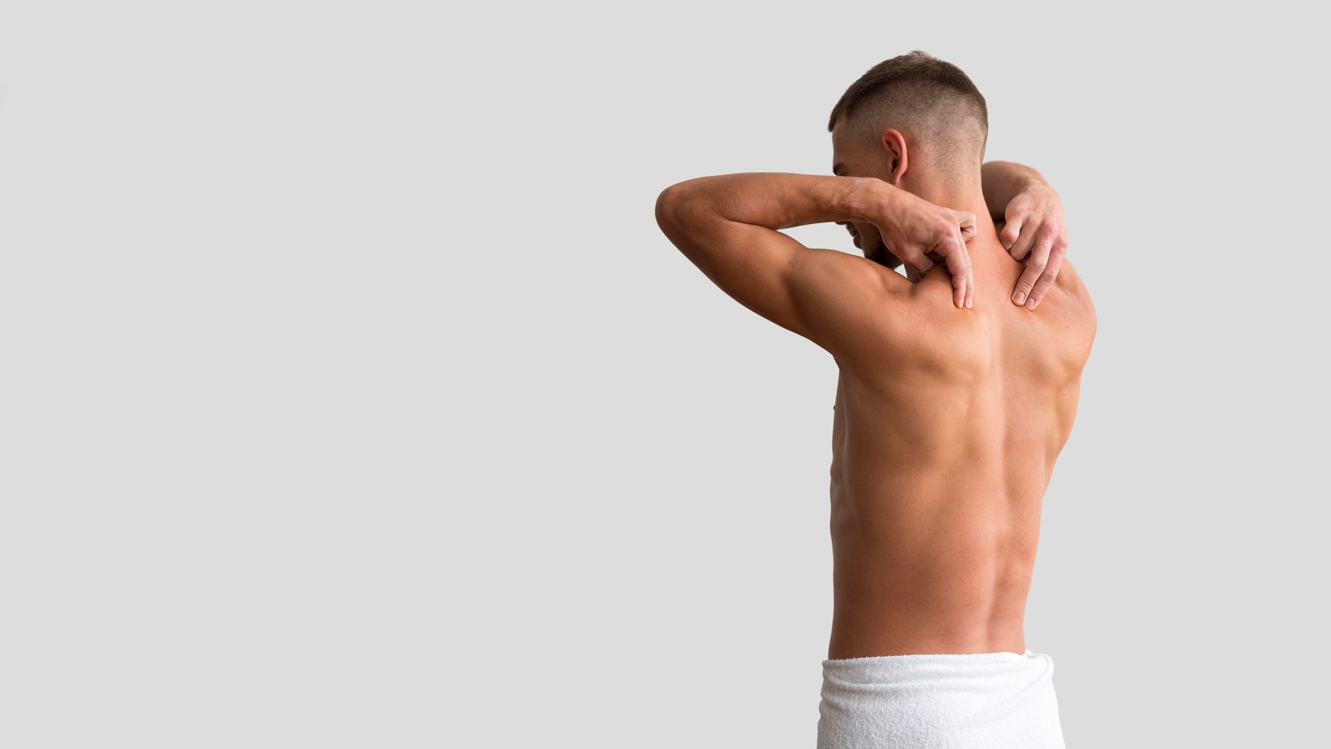 Man shirtless with towel around waist, stretching arms behind head against a plain light background