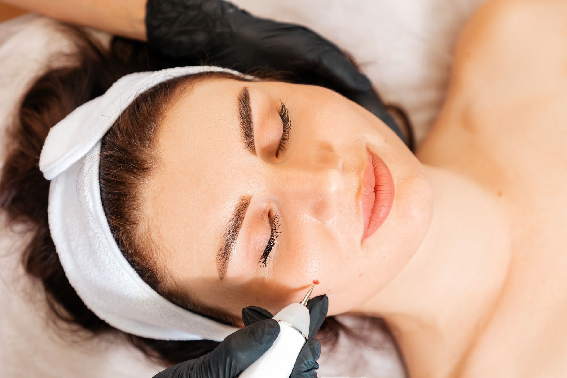 Person receiving facial treatment with a cosmetic tool, eyes closed, wearing a white headband