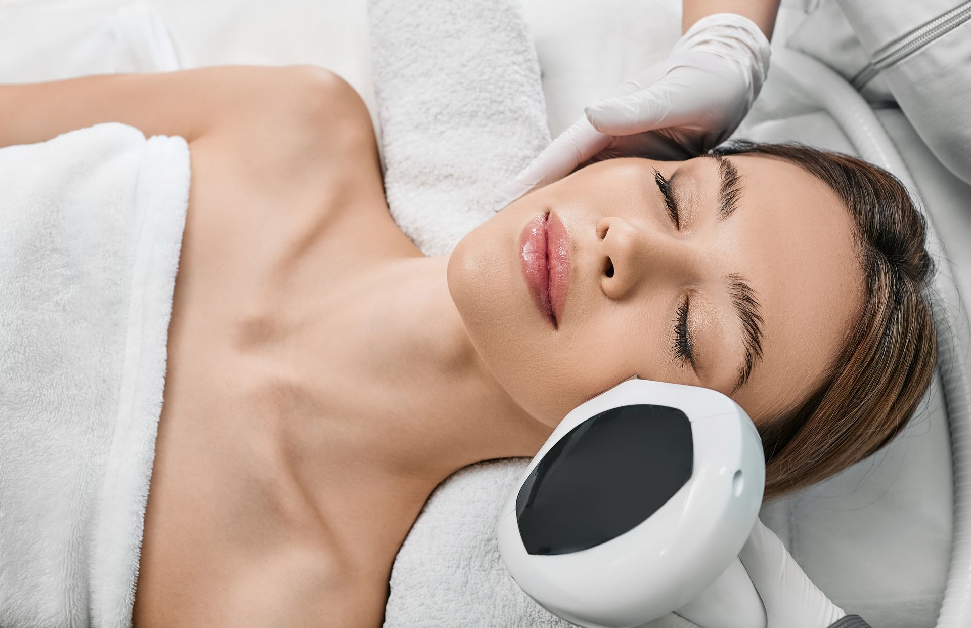 Woman receiving facial treatment, lying on white towel, technician using a device on her face.