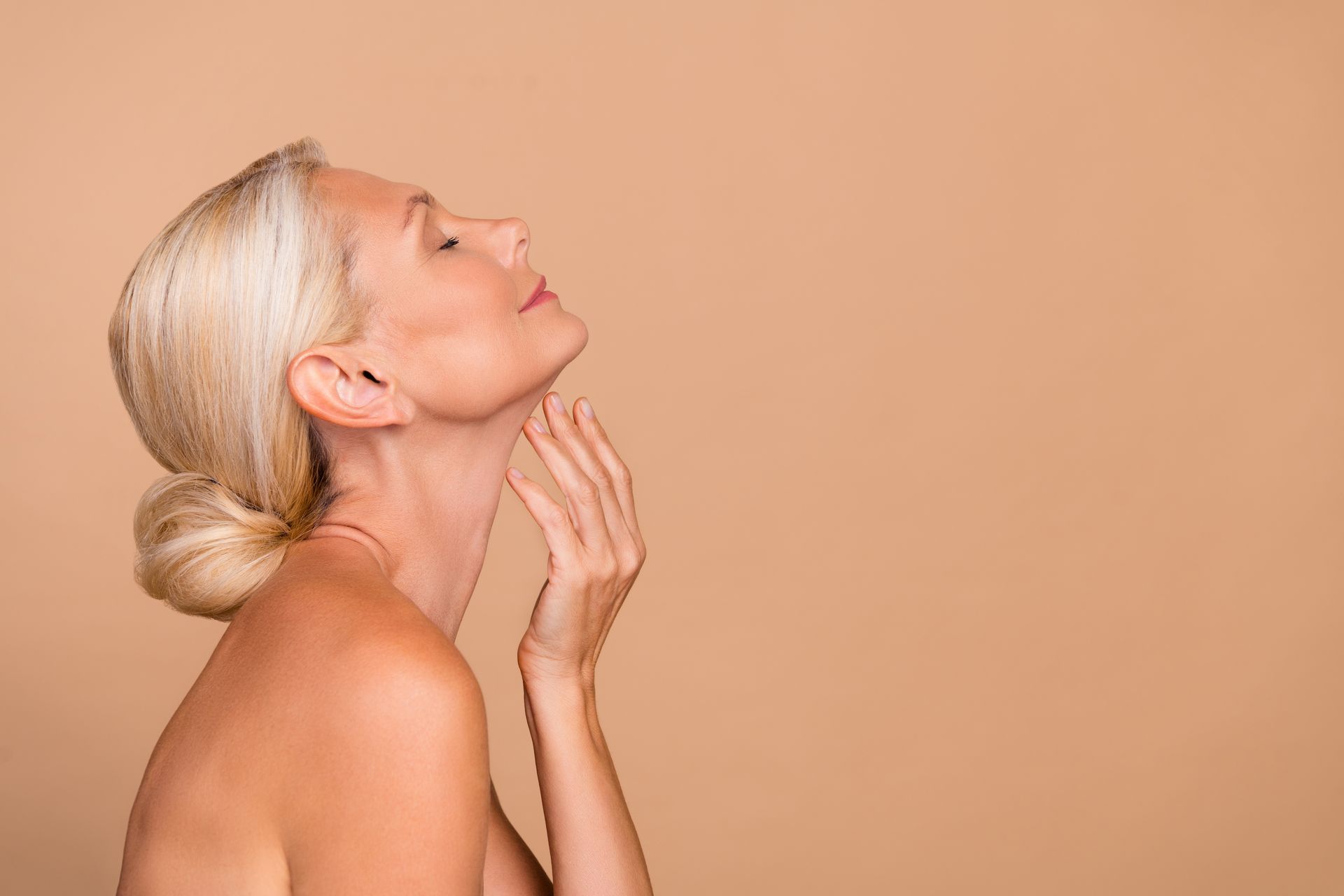 Blonde woman with bun applying cream to her neck against a peach background