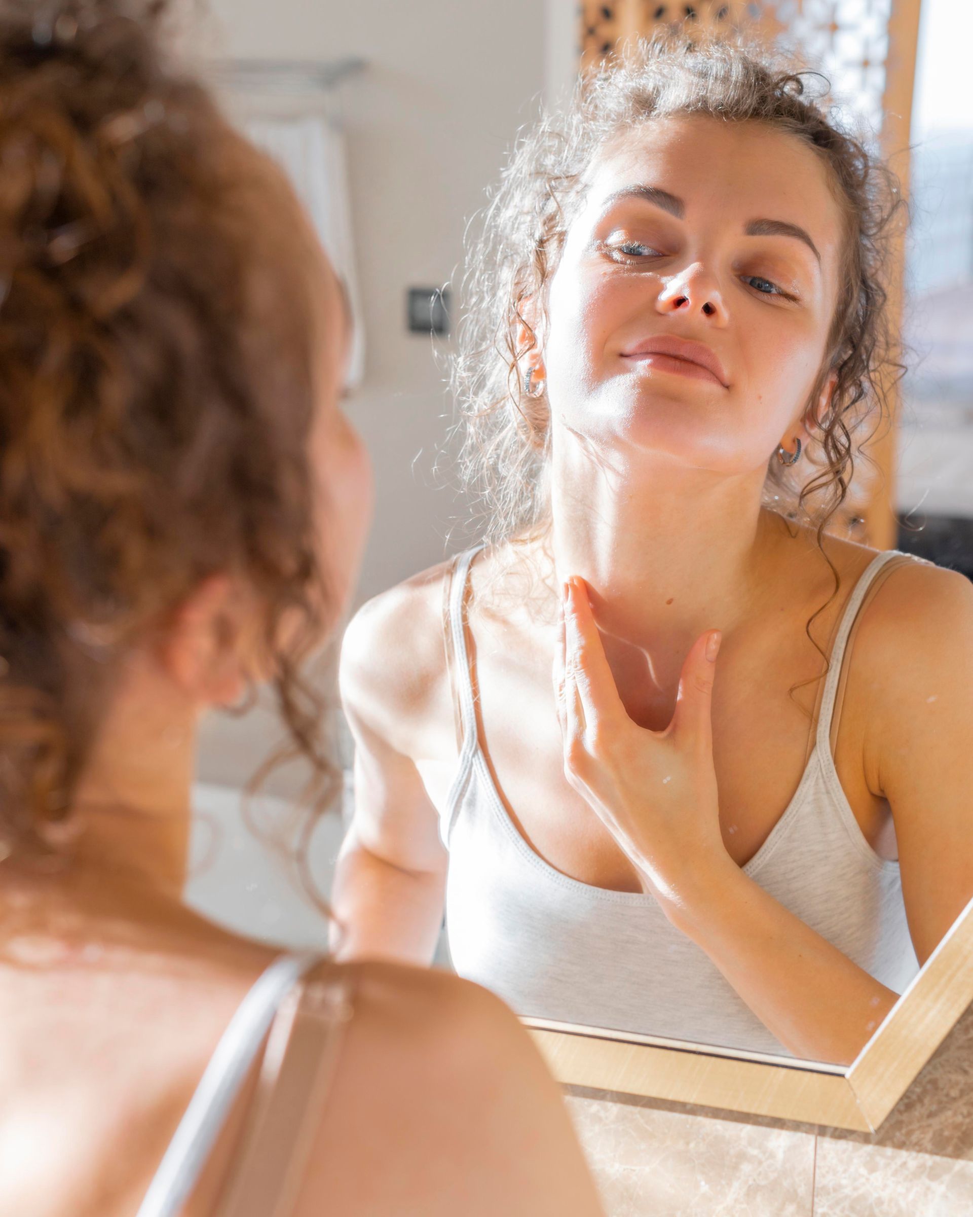 Woman applying skincare to her neck while looking in a mirror in a bright bathroom