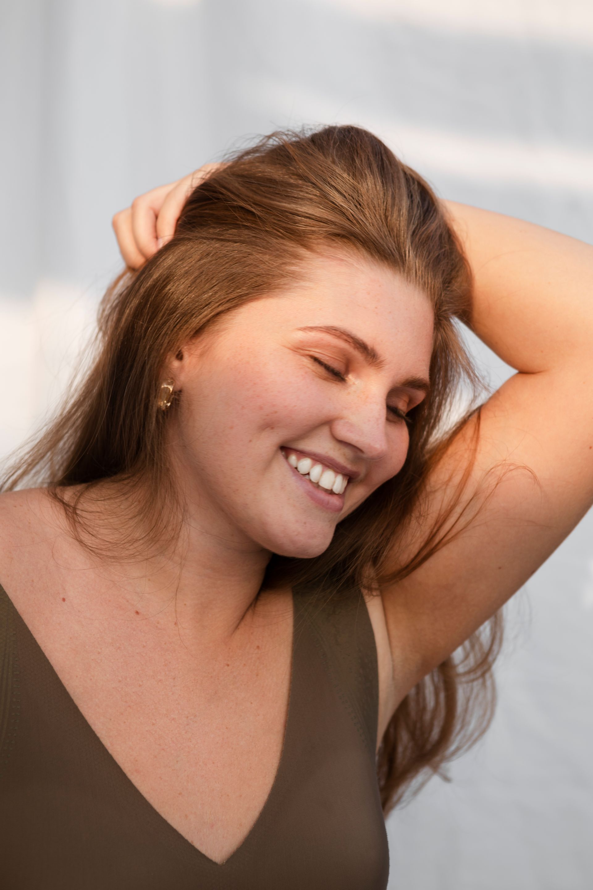 Smiling woman with long brown hair, eyes closed, stretching with one arm behind her head indoors