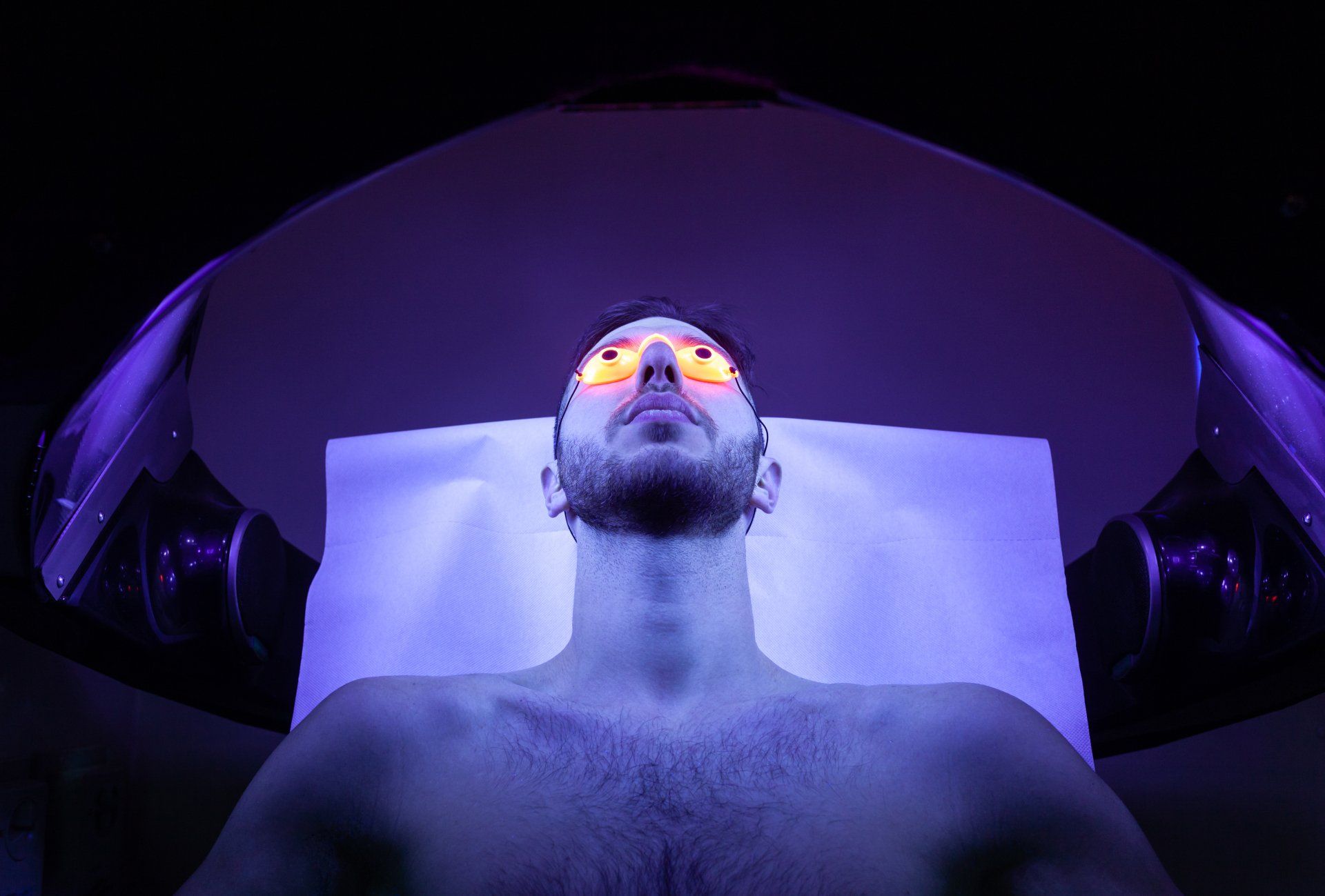 Man lying under purple lighting with glowing orange goggles, viewed from below in a dark room