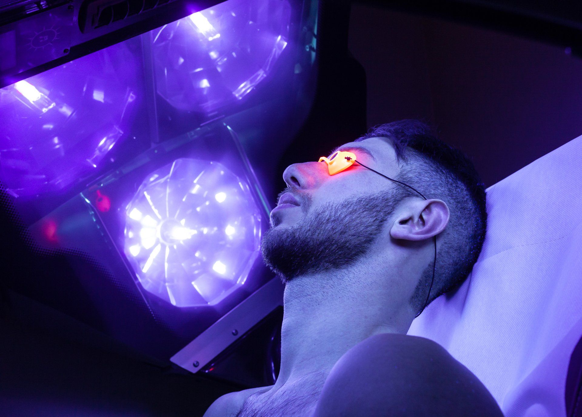 Man under purple LED lights wearing glowing red glasses, reclining on a white surface
