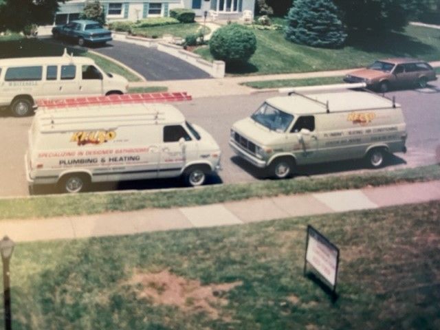 Two plumbing vans parked on a residential street. Both are white with company logos.