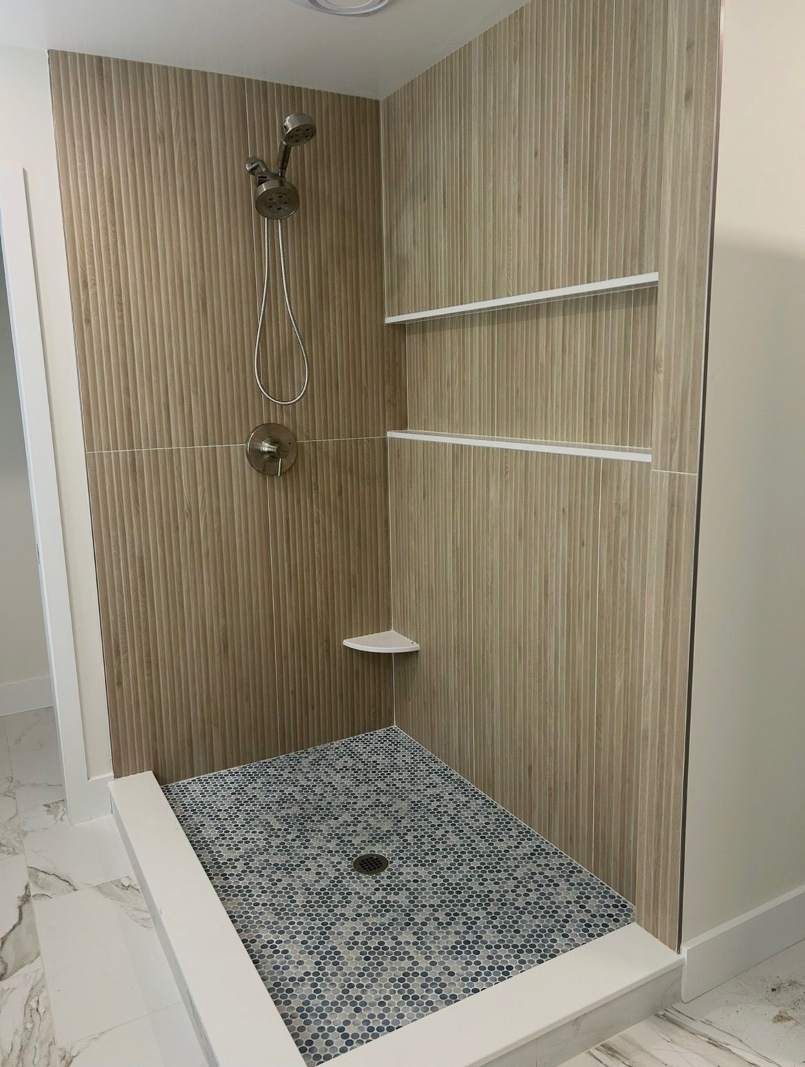 Corner walk-in shower with beige tile walls, gray mosaic floor, chrome showerhead, and small shelf.