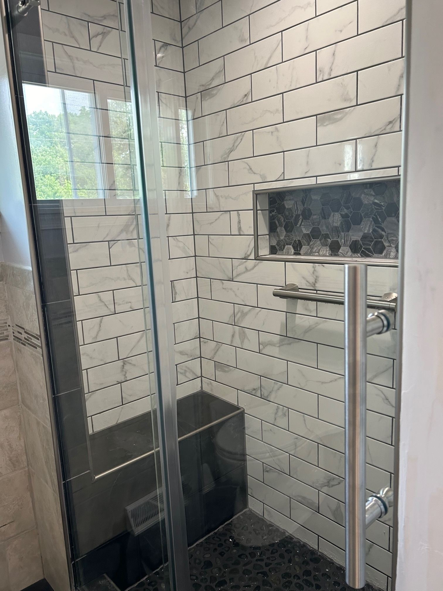 Modern tiled shower with glass door, wall-mounted handheld shower, and black bench.