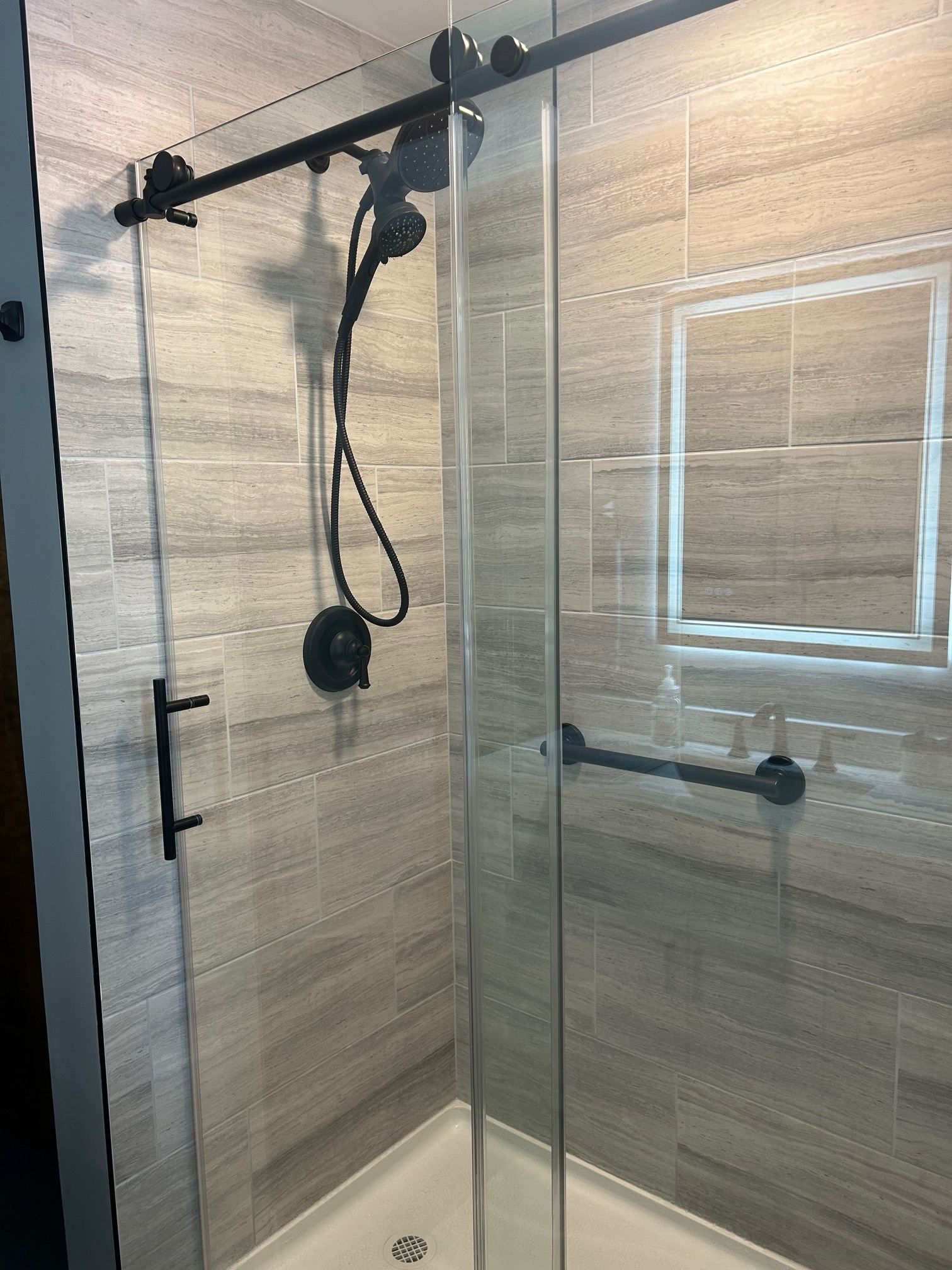 Walk-in shower with black showerhead and controls, beige tile walls, and glass door with metal handle