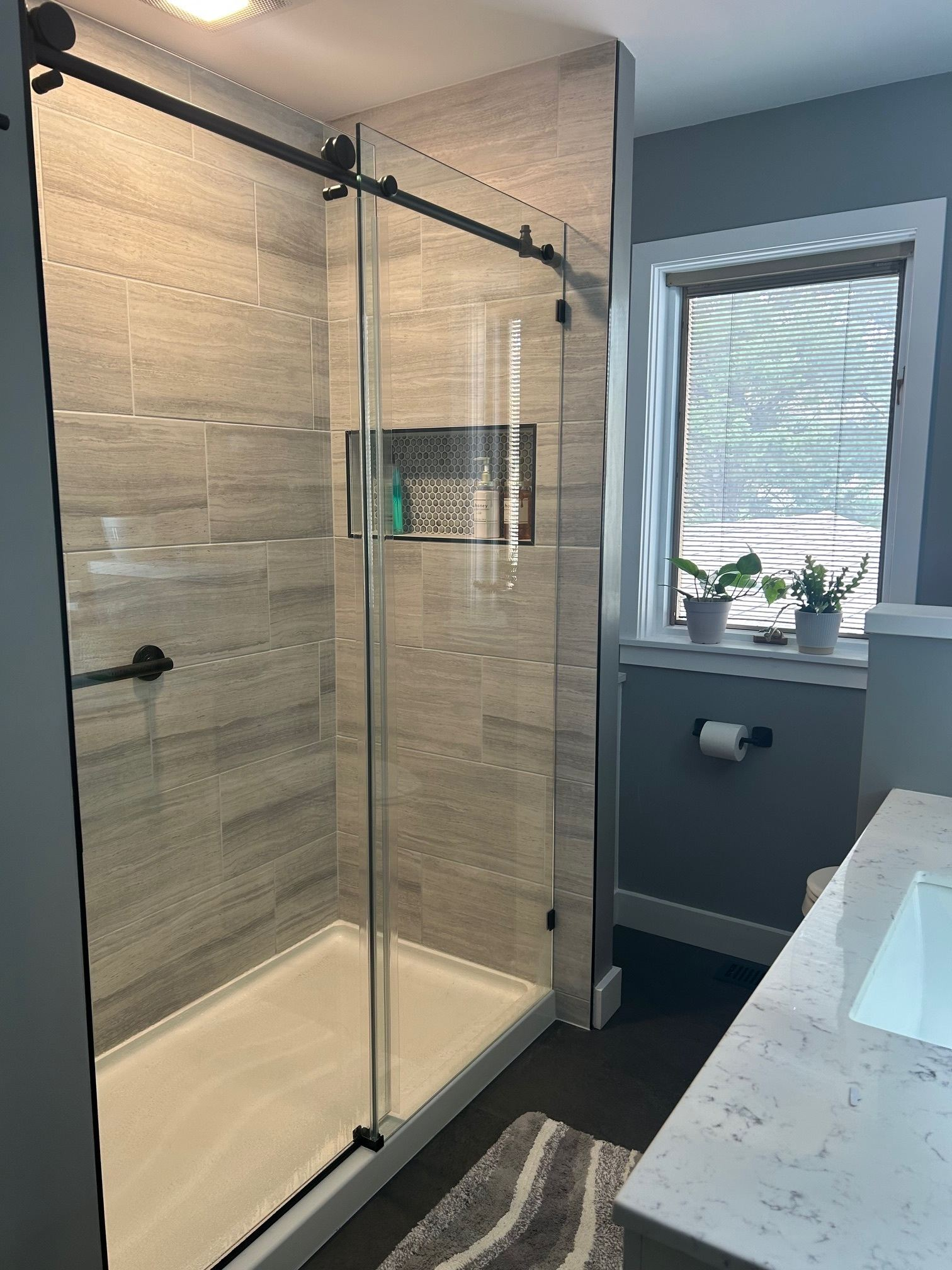 Modern bathroom with glass shower enclosure, gray tile walls, and window light.