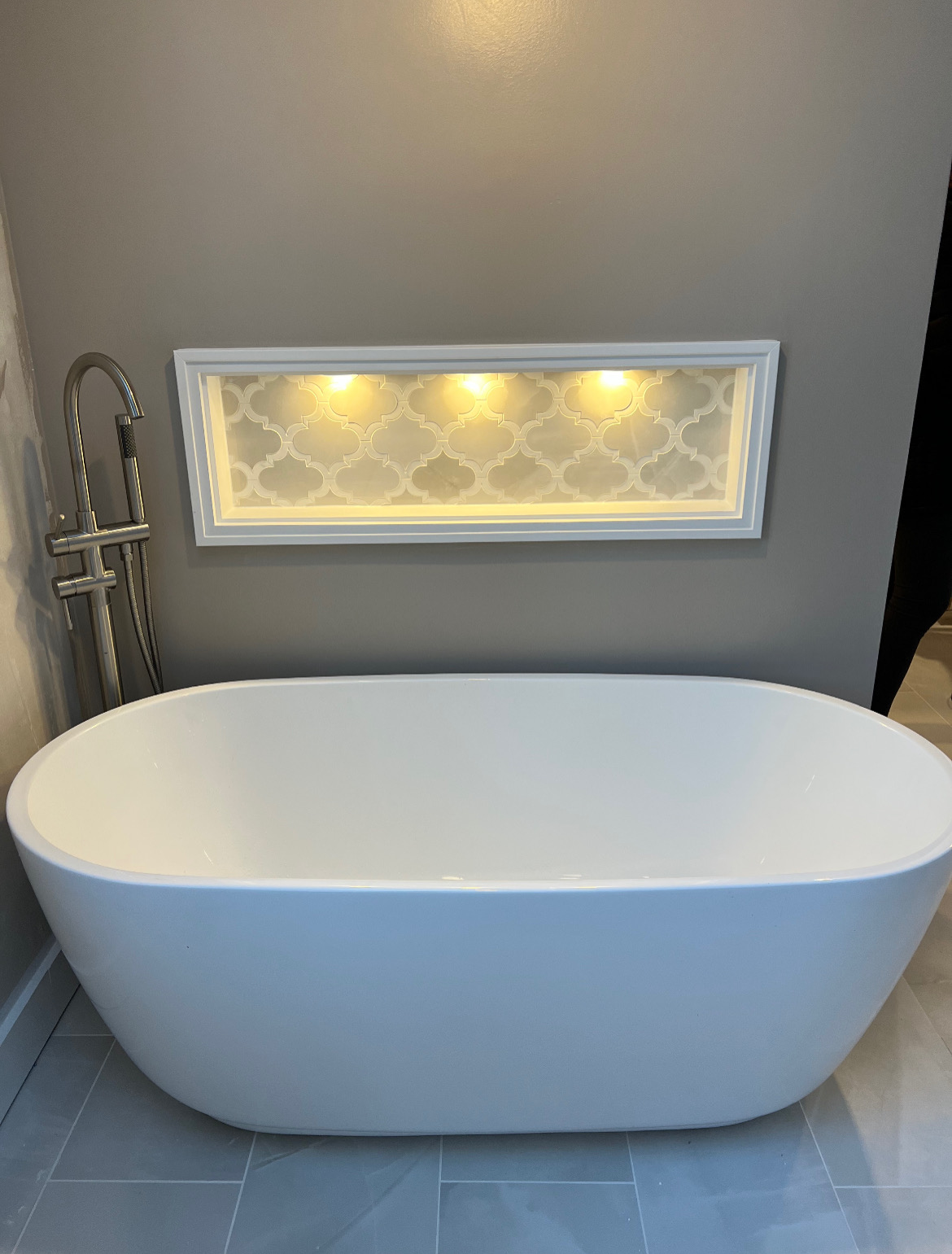 Modern white freestanding bathtub beneath a backlit niche in a gray-tiled bathroom