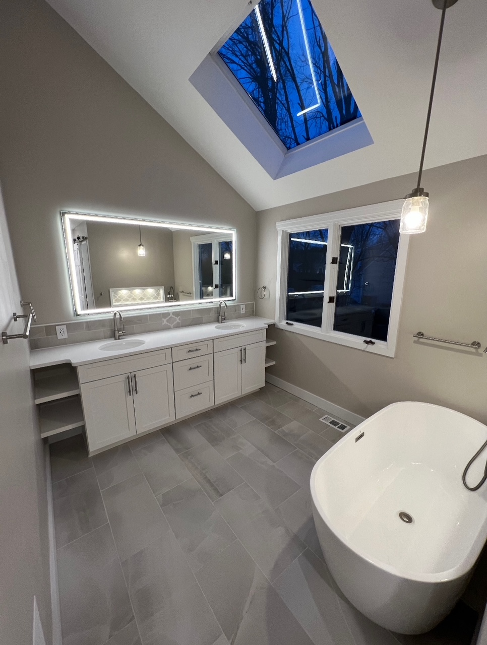 Modern bathroom with skylight, large mirror, vanity, and freestanding bathtub on gray tile floor