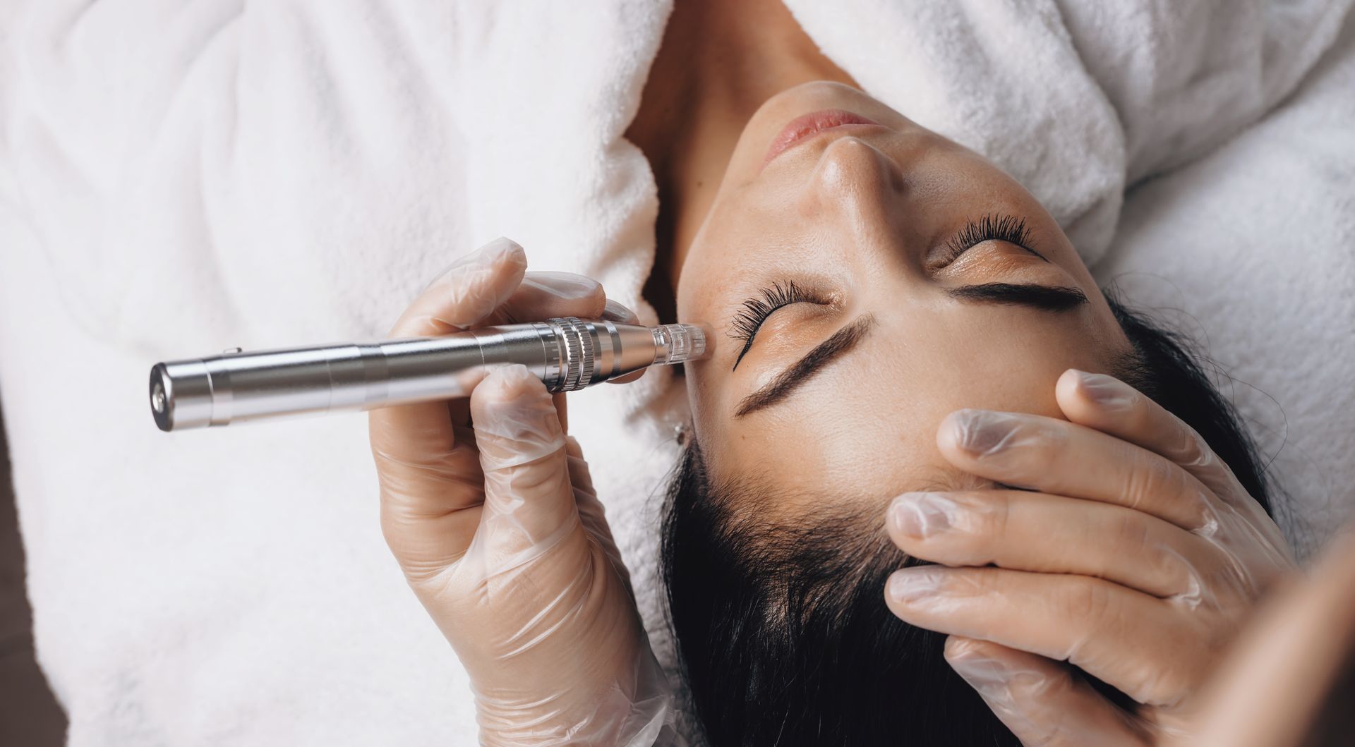 Spa treatment with a woman receiving a facial massage using a handheld device, eyes closed and resting on a towel.