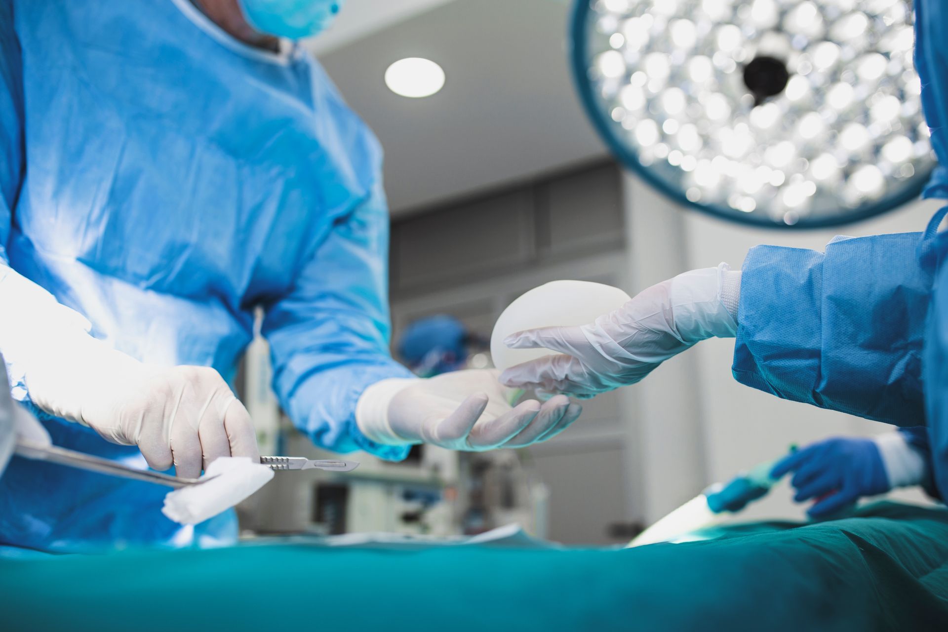 Surgeons in blue scrubs and gloves passing an instrument in an operating room under bright surgical lights