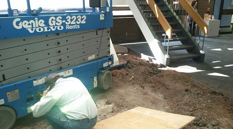A person inspects a blue Genie lift next to stairs and dirt in a building.