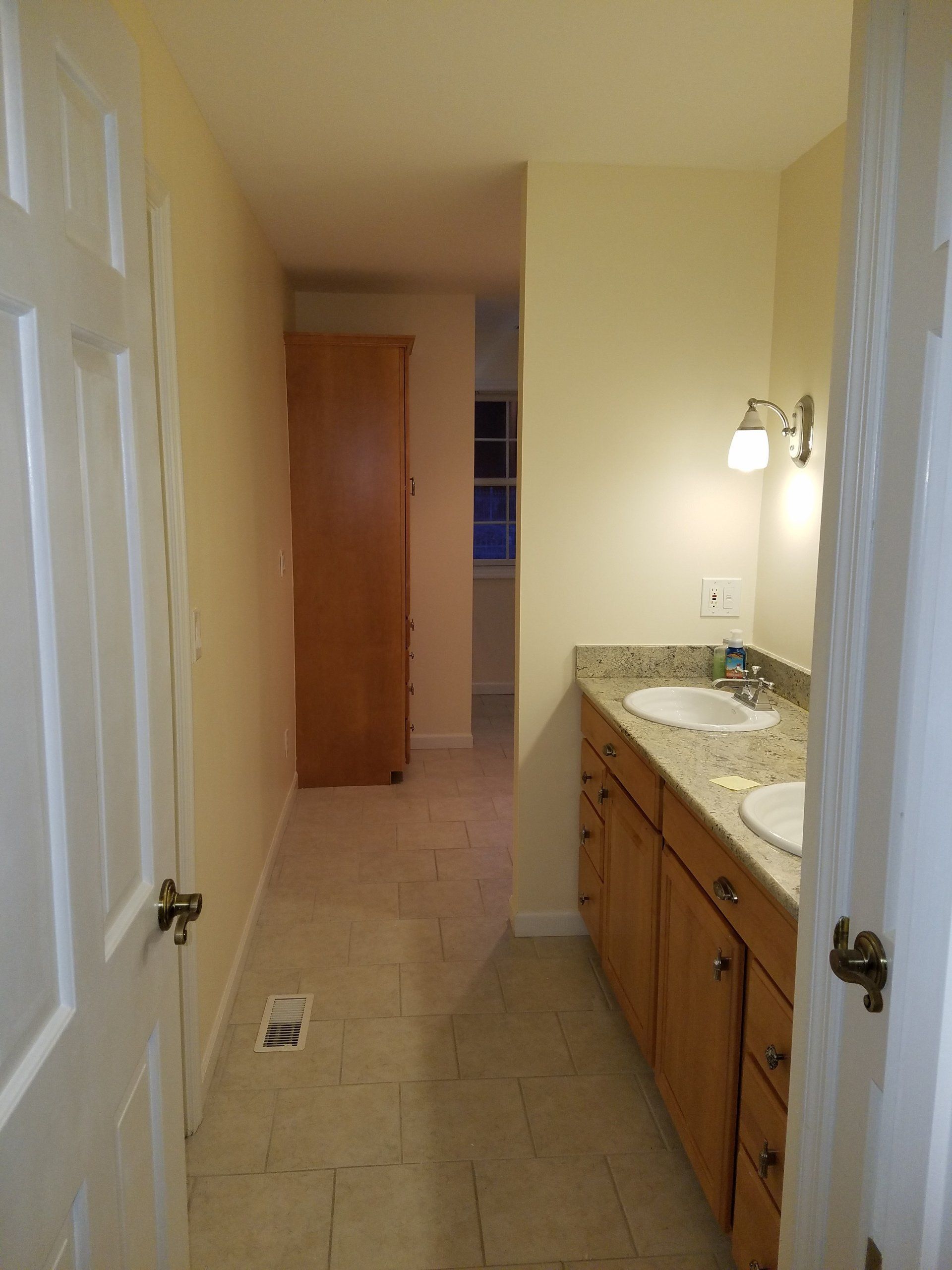 Bathroom with light-colored walls, vanity with two sinks, and a tall cabinet in the hallway.